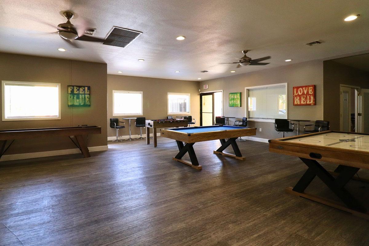 A spacious game room featuring a wooden air hockey table, a foosball table, and colorful wall art with motivational phrases. The room has large windows for natural light and a modern aesthetic, including ceiling fans and contemporary bar-style seating.