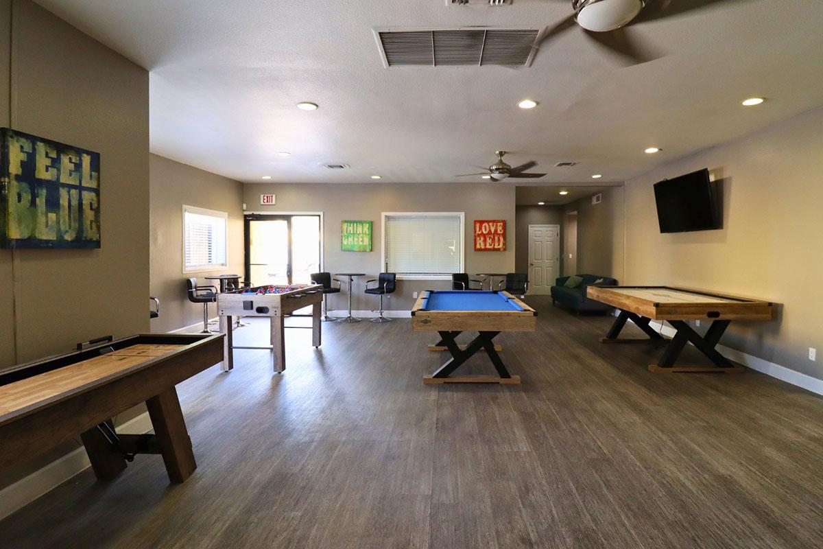 An indoor recreational area featuring a pool table, foosball table, and two wooden game tables. The walls are decorated with colorful motivational posters. There are chairs and a sofa, with large windows allowing natural light to fill the space. A flat-screen TV is mounted on the wall.
