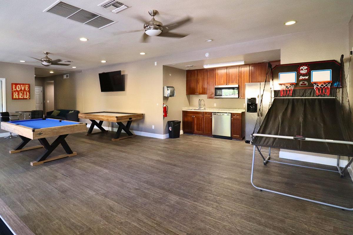 Interior of a recreational room featuring a blue pool table, a basketball shooting game, and a kitchenette with wooden cabinets. There is a television on the wall and a seating area in the background, all set in a modern design with wood flooring.