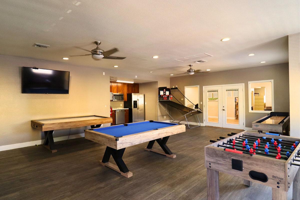 Recreational game room featuring a pool table, foosball table, and air hockey table. The space includes a large flat-screen TV, modern kitchen area with appliances, and a warm, inviting atmosphere with ceiling fans and wooden flooring. Brightly lit with an open layout.