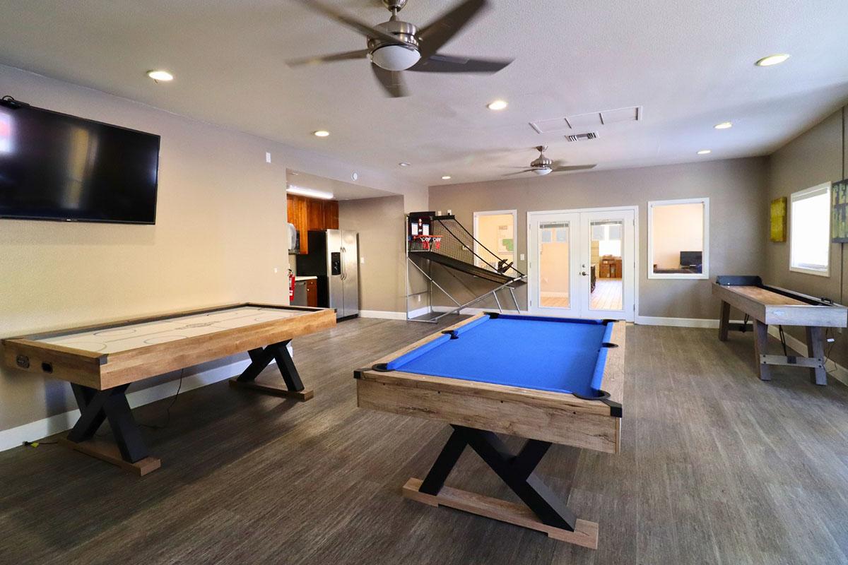 A recreational room featuring a blue pool table, a wooden air hockey table, and a table for games. There is a wall-mounted television, large windows allowing natural light, and a modern kitchen visible in the background. The decor is simple and inviting, with a polished wooden floor.