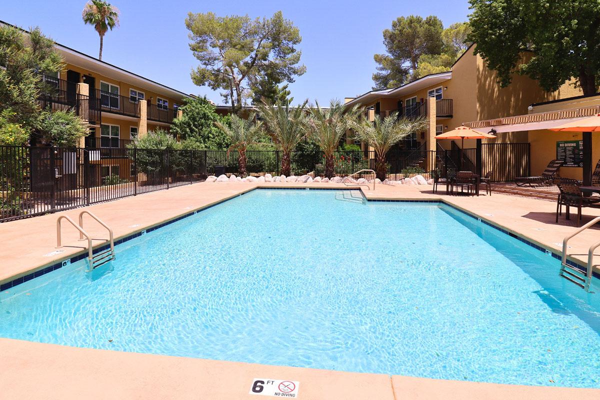 A clear blue swimming pool surrounded by palm trees and lounge chairs, with two sets of steps leading into the water. In the background, buildings with balconies are visible, and the scene is set in a sunny outdoor environment. A "6 ft" sign is visible near the pool edge.