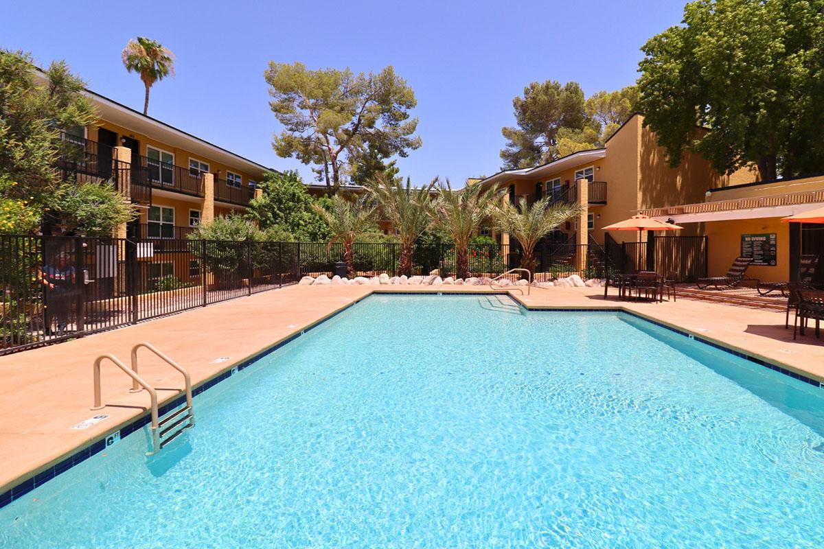 A bright outdoor swimming pool surrounded by palm trees and green landscaping. In the background, there are several multi-story apartment buildings with balconies. The pool area features lounge chairs and orange umbrellas, creating a relaxing atmosphere on a sunny day.