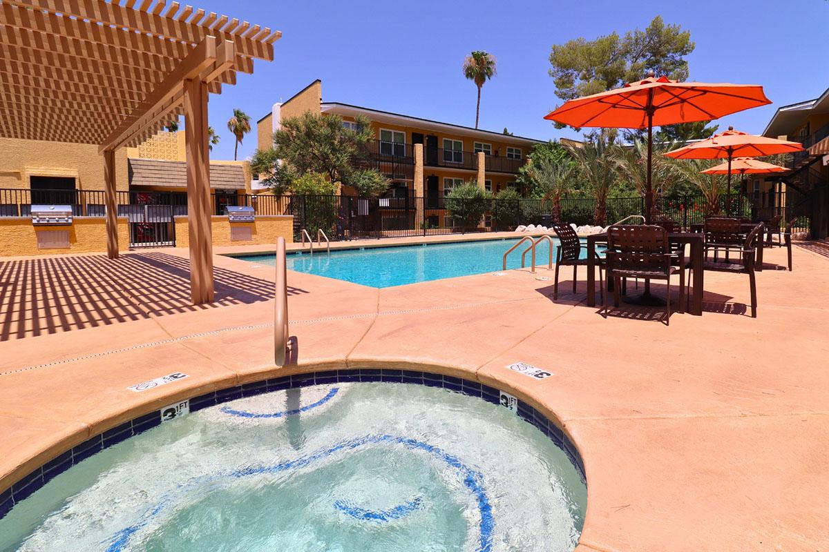 A sunny pool area featuring a sparkling blue swimming pool surrounded by lounge chairs and orange umbrellas. A connected hot tub is visible in the foreground, with a backdrop of palm trees and apartment buildings. The scene captures a relaxing outdoor atmosphere ideal for leisure.