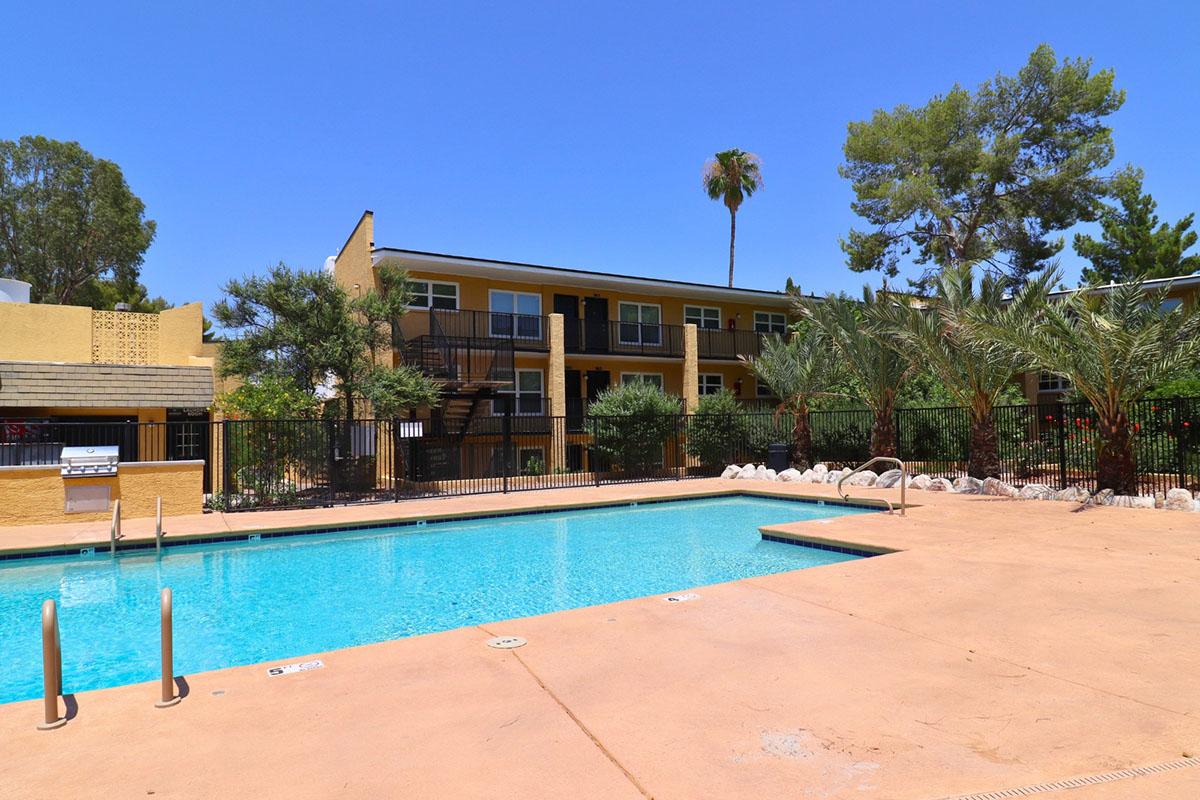 A clear swimming pool surrounded by a patio area with lounge chairs. In the background, there are two-story apartment buildings and palm trees, set against a bright blue sky. The area is fenced with greenery and a barbecue grill nearby, creating a relaxing outdoor space.