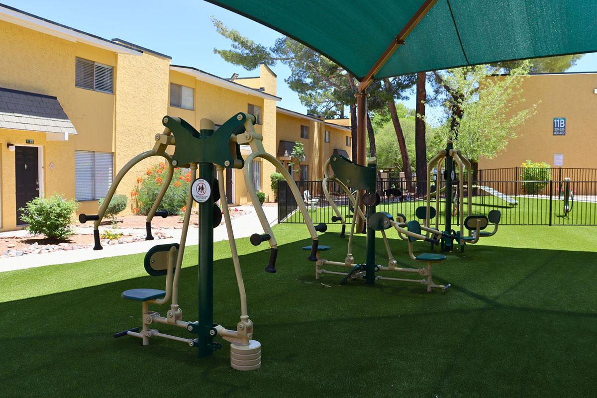 Outdoor fitness area featuring several exercise machines under a green canopy, set on artificial grass. Apartment buildings are visible in the background, surrounded by trees. The space is designed for community use, promoting health and wellness.
