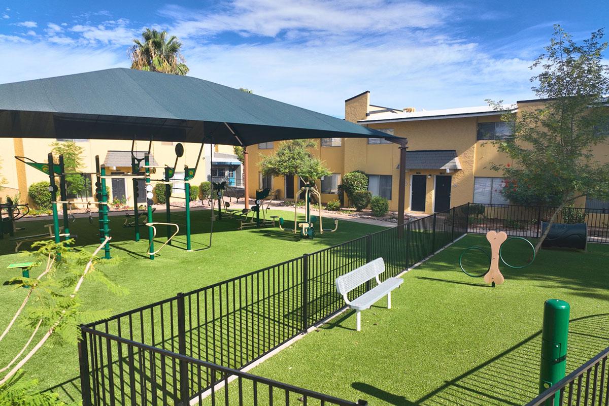 Playground area featuring various outdoor exercise equipment under a shaded canopy, surrounded by green artificial grass. There are benches for seating, pathways, and a few trees, with residential buildings in the background against a clear blue sky.