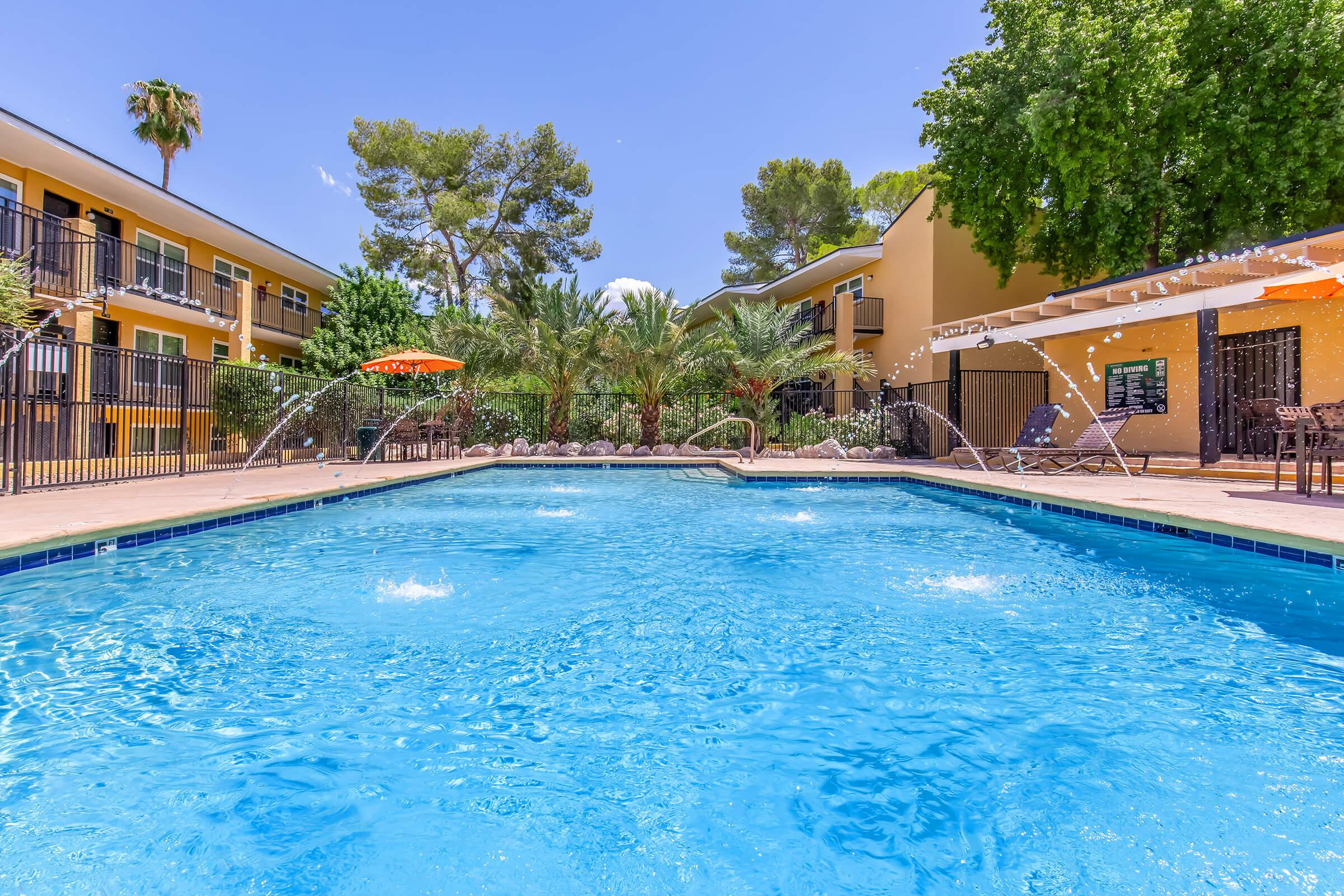 A vibrant outdoor pool surrounded by lush greenery and palm trees, with water features spraying into the air. Comfortable lounge chairs and shaded areas with umbrellas are visible. The setting is bright and inviting, suggesting a relaxing atmosphere for leisure and enjoyment.