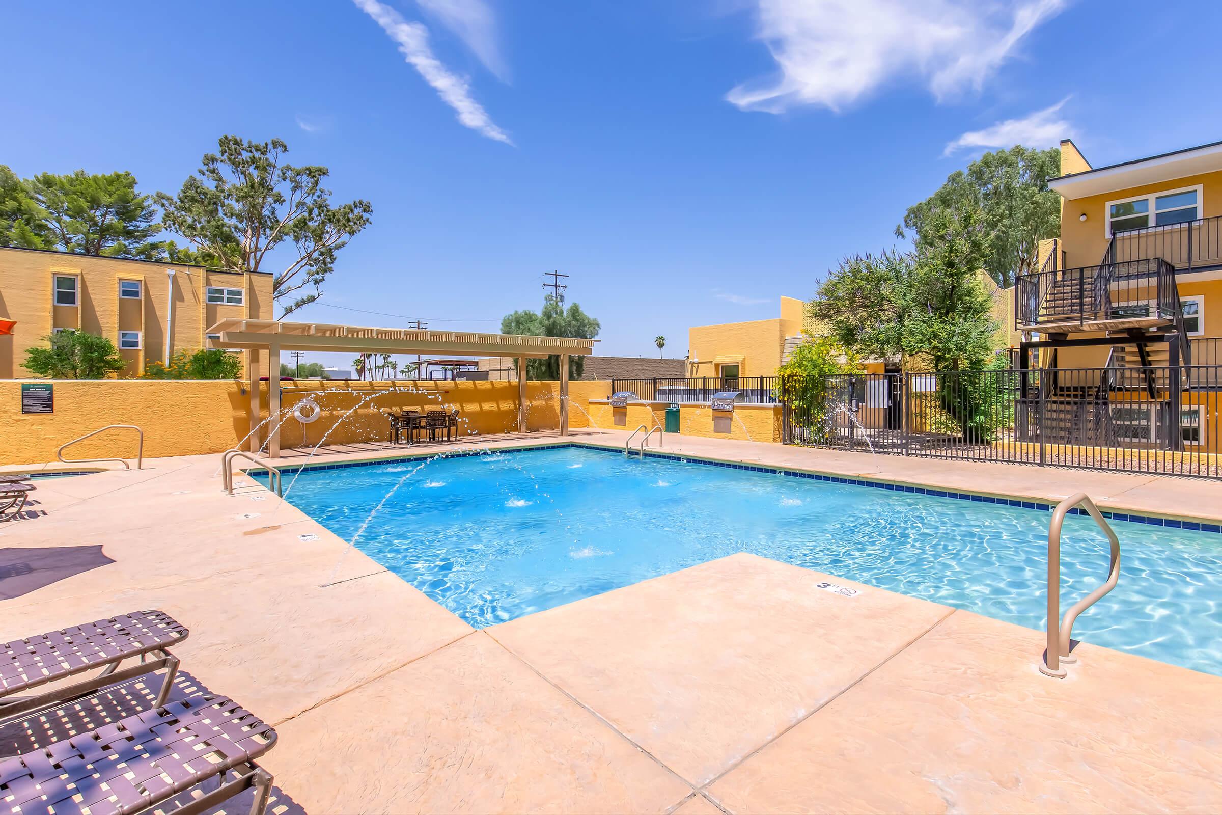A clear blue swimming pool surrounded by lounge chairs, palm trees, and a sunny sky. In the background, there are buildings with a warm yellow hue and a shaded seating area. The scene conveys a vibrant and inviting atmosphere perfect for relaxation.