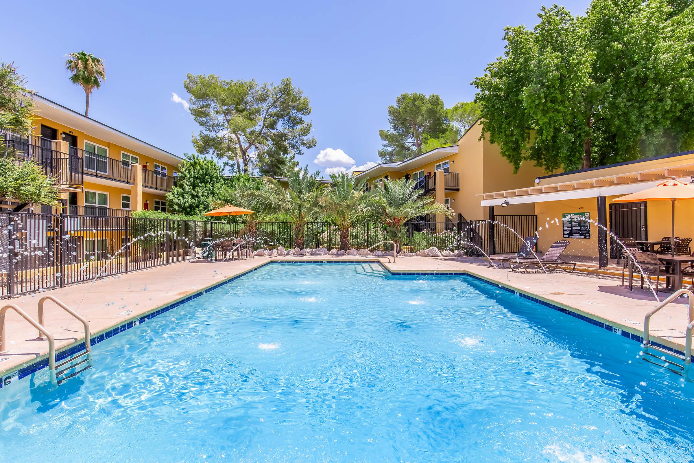 A clear blue swimming pool surrounded by lounge chairs, palm trees, and orange umbrellas. Apartment buildings with yellow exteriors are visible in the background under a bright blue sky with a few clouds. The scene conveys a relaxing outdoor atmosphere.
