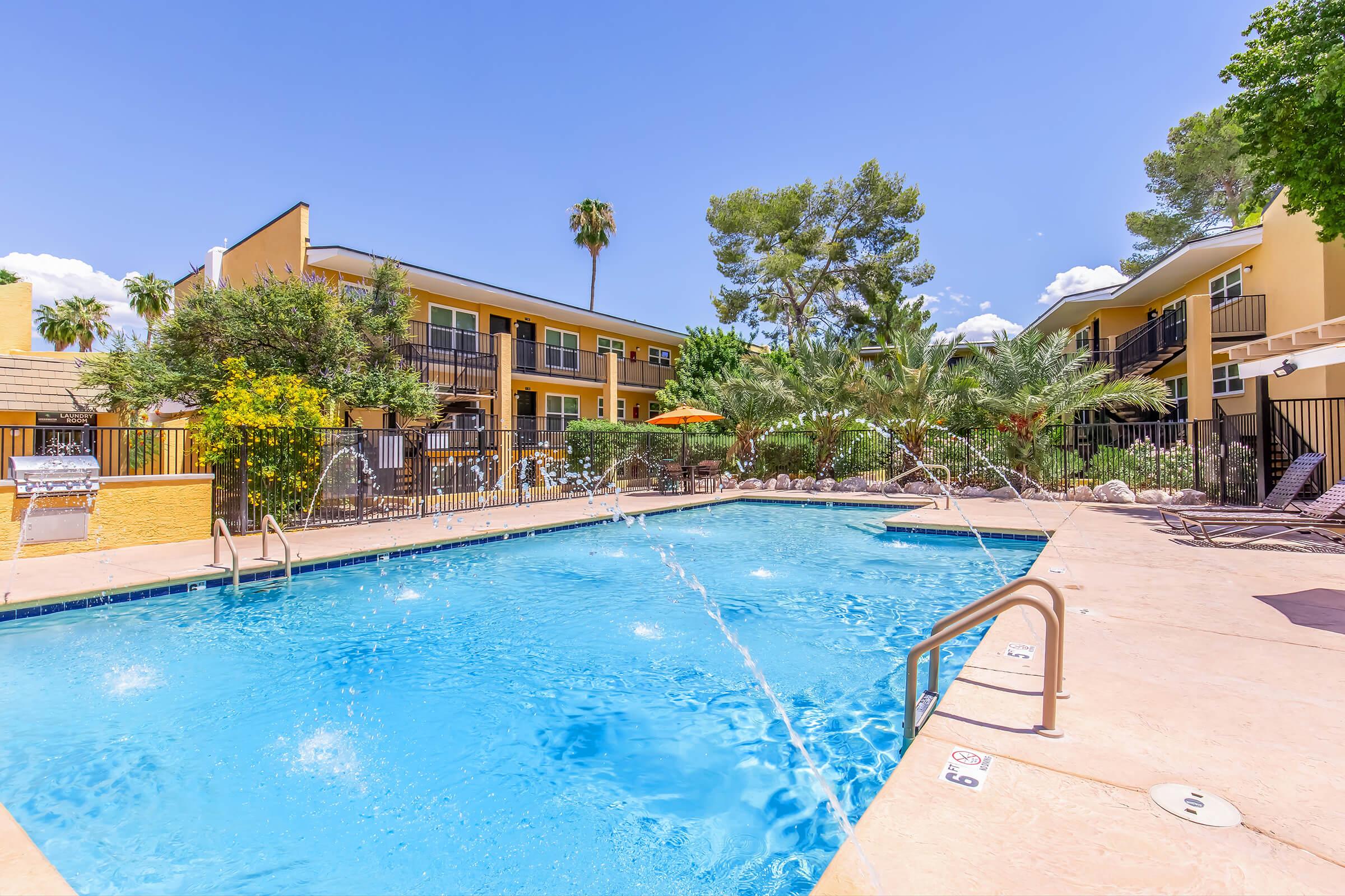 A vibrant outdoor swimming pool surrounded by lush greenery and palm trees. The pool area features lounge chairs, a splash fountain, and two-story yellow apartment buildings in the background under a clear blue sky. Brightly colored umbrellas provide shade near the poolside.