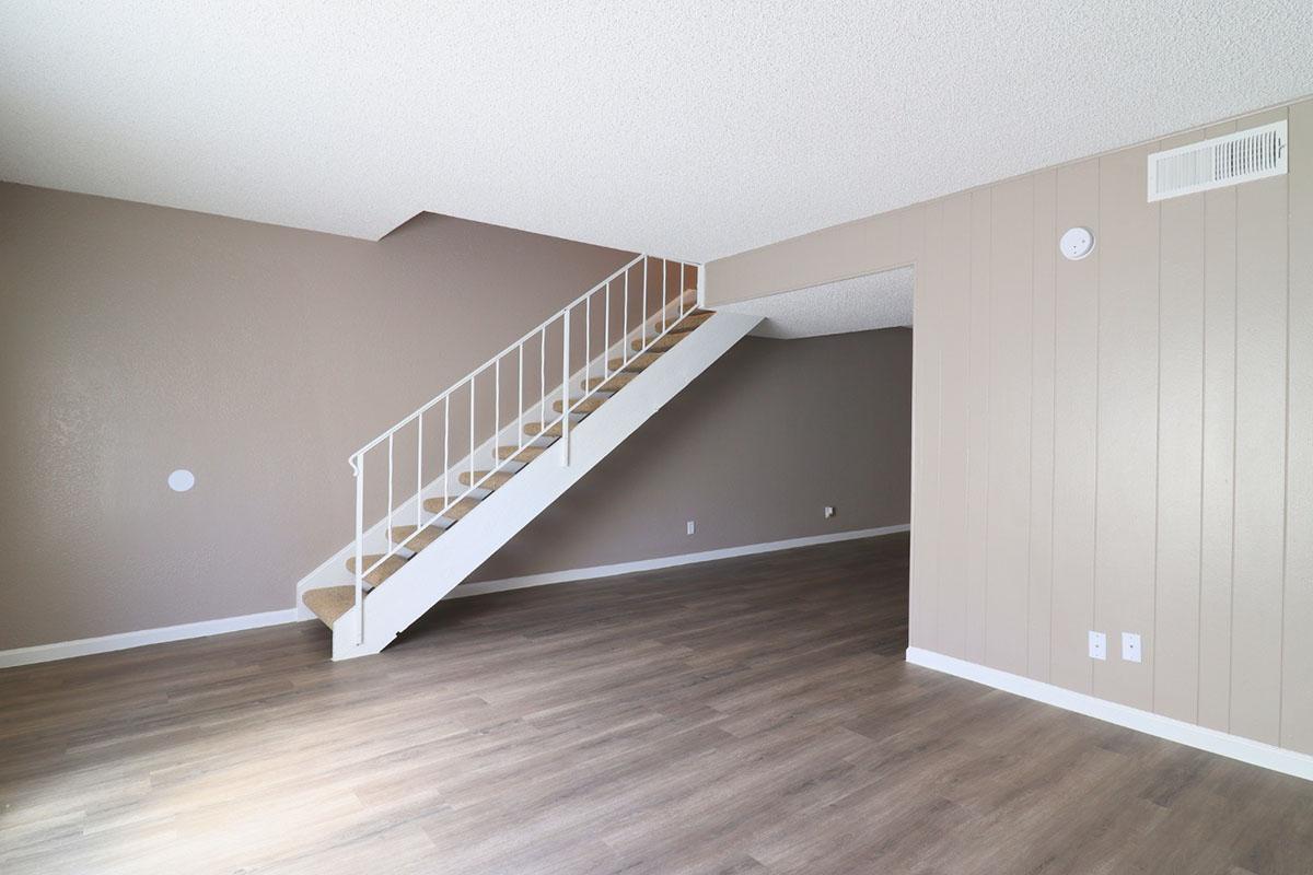 A spacious, empty living room with light brown walls and laminate flooring. A staircase with a white railing leads to the upper level. The room features a simple design, with a small nook visible on one wall and no furniture, creating an open and airy atmosphere.