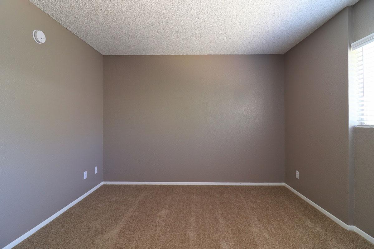 A vacant room with beige carpeting and light brown textured walls. There is a smoke detector on the ceiling and a window with blinds partially visible on the right side. The room has no furniture, creating an open and spacious feel.