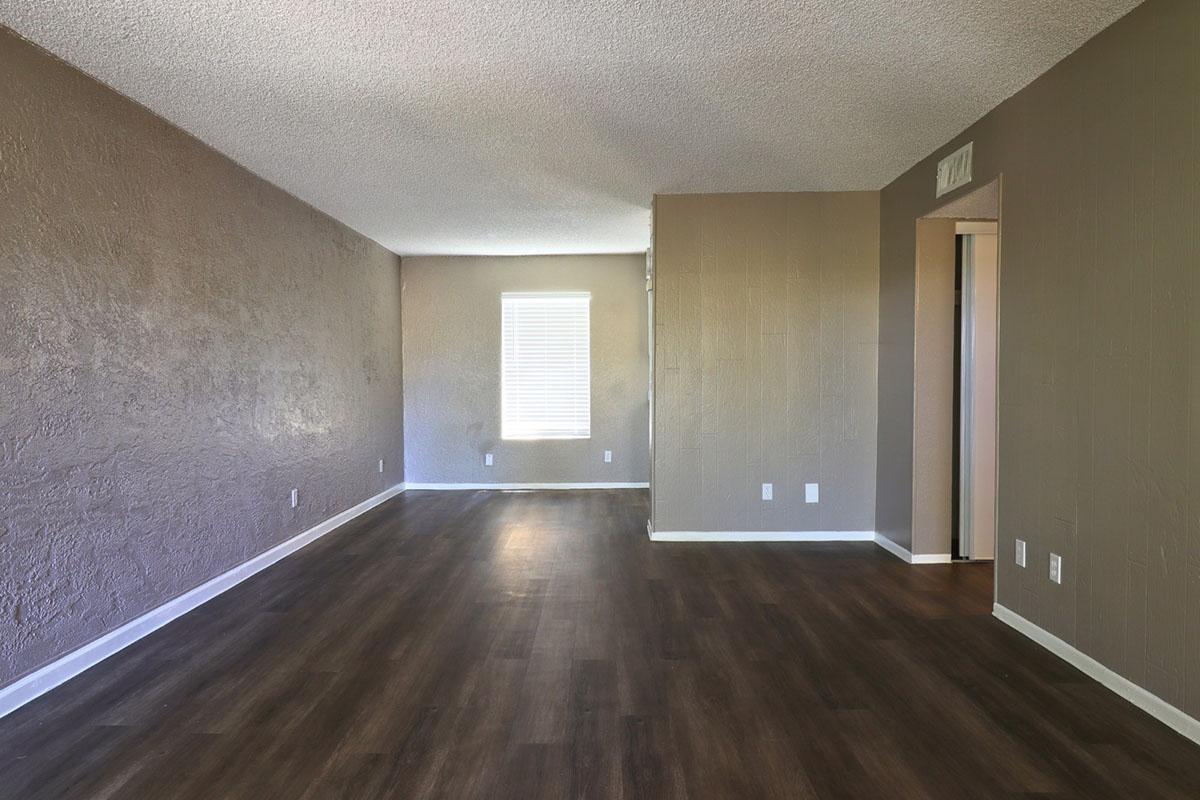 A spacious, empty room with textured brown walls and light-colored wooden flooring. A window on one wall allows natural light to enter, while another wall features an opening leading to a hallway. The overall atmosphere is clean and ready for decoration or new furniture.