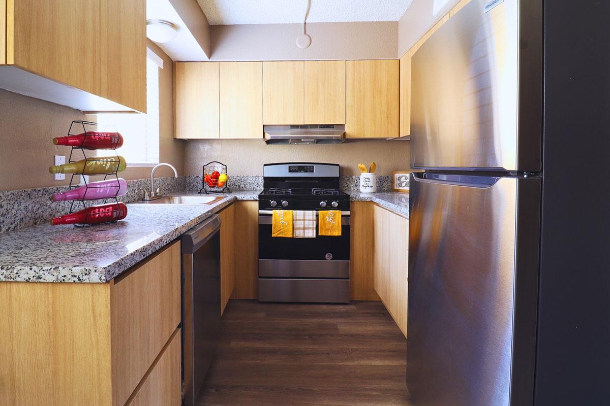 Modern kitchen featuring light wood cabinetry, a stainless steel refrigerator, a gas stove with an oven, and dark granite countertops. The space includes a dishwasher, colorful kitchenware, and a fruit bowl, with natural light coming in through a window. The flooring is wood-like laminate.