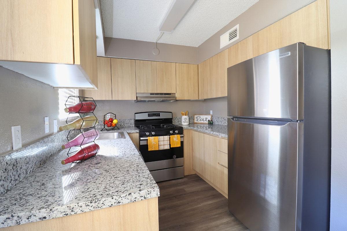 A modern kitchen featuring light wood cabinetry, a stainless steel refrigerator, and a granite countertop. It includes a gas stove, a black wire fruit basket with apples and oranges, and a decorative towel hanging on the oven. The overall design is clean and contemporary with neutral tones.