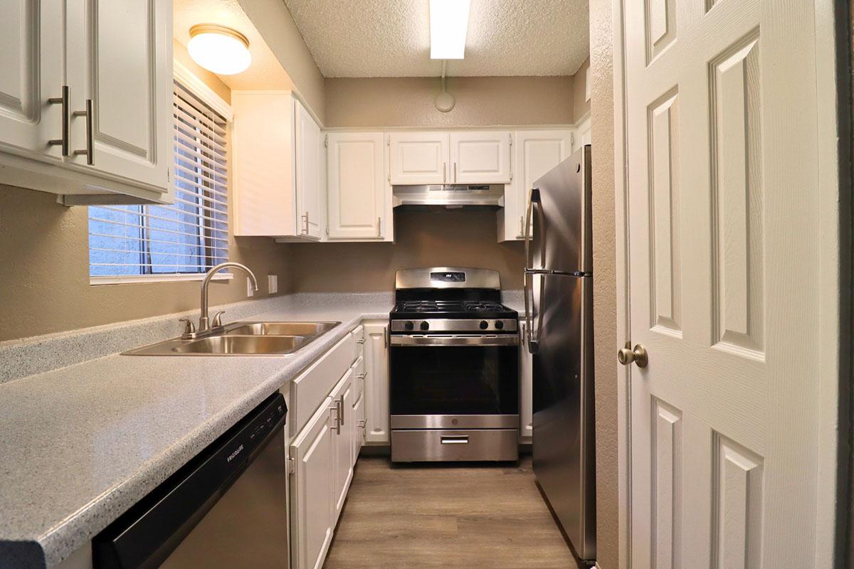 A modern kitchen featuring white cabinetry, a stainless steel stove and refrigerator, a double sink, and granite countertops. There is a window providing natural light and a light fixture above the sink. The flooring is a light wood laminate, enhancing the space's contemporary feel.