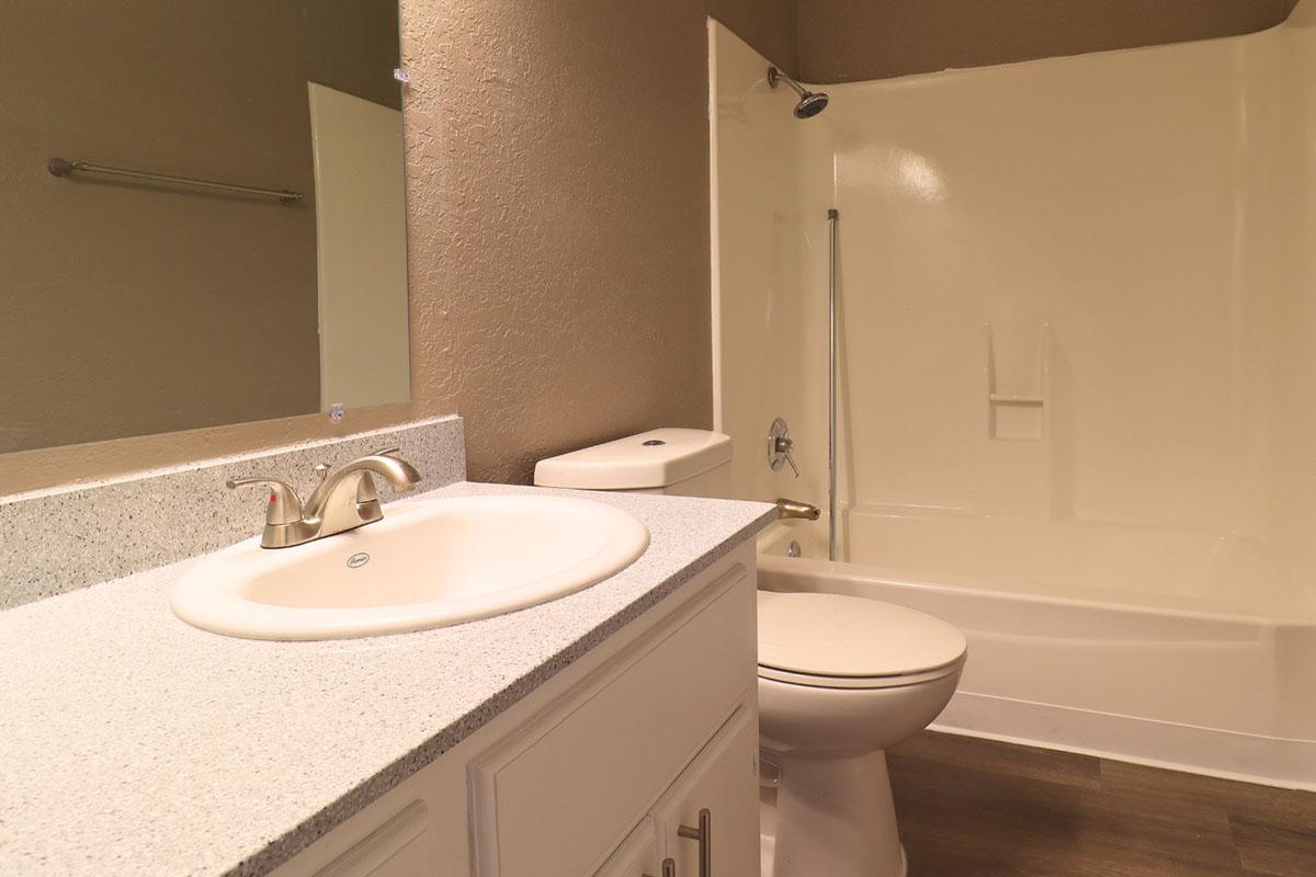 A clean and modern bathroom featuring a white countertop with a sink and faucet, a toilet, and a shower-tub combination. The walls are painted light brown, and a mirror is mounted above the sink. The flooring is a dark wood, enhancing the contemporary look of the space.