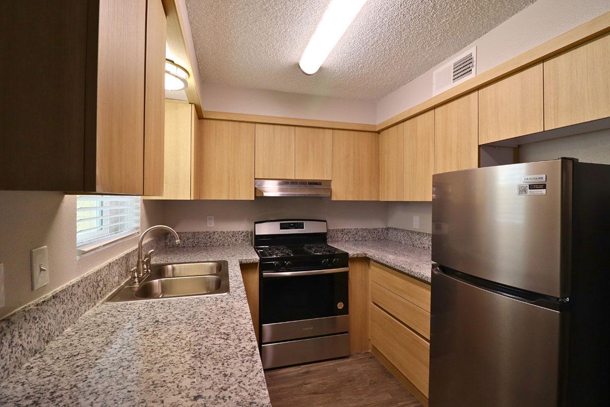 Modern kitchen with light wood cabinetry, stainless steel appliances, including a refrigerator and stove, and a granite countertop. The space features a double sink, overhead lighting, and windows that provide natural light. The overall design is clean and contemporary.