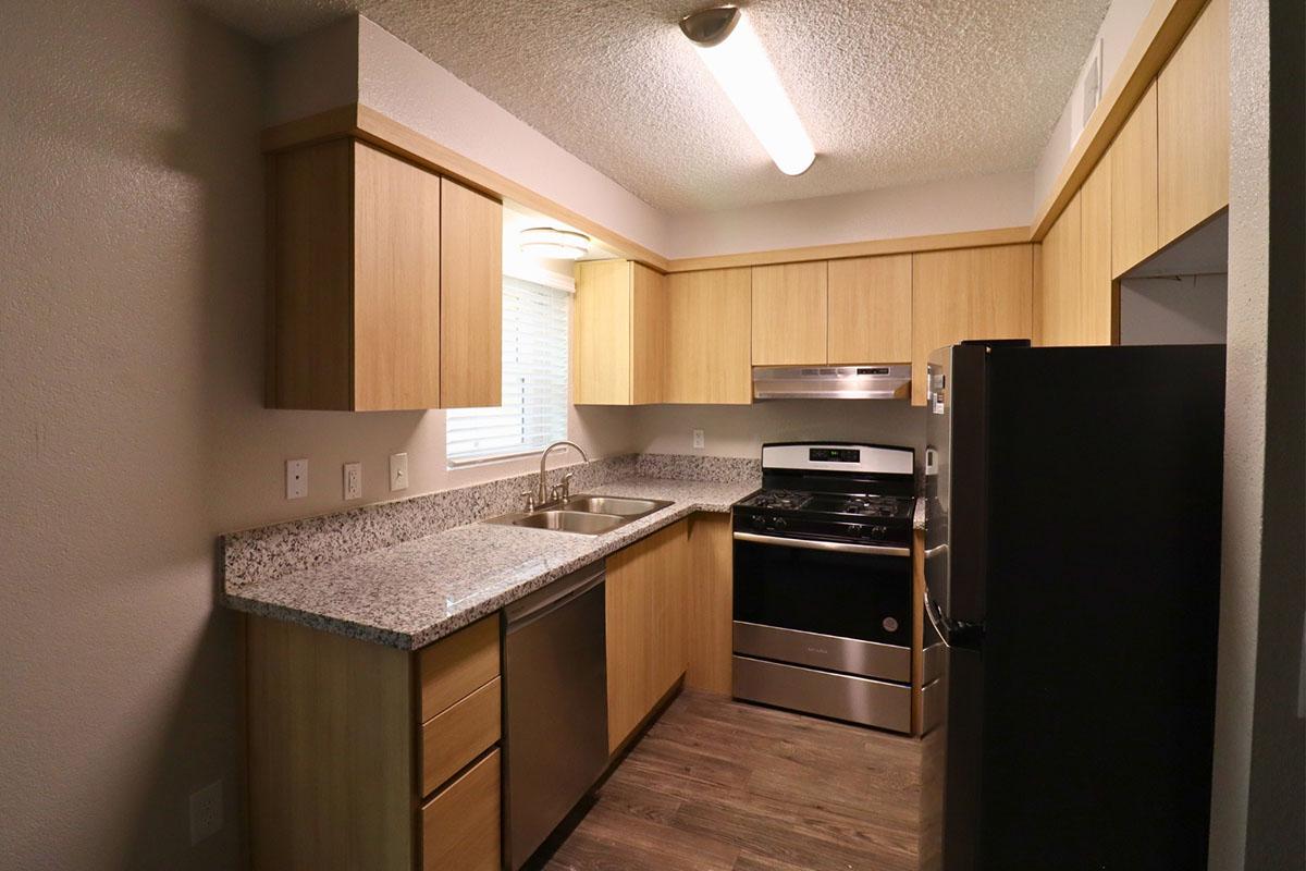 A modern kitchen featuring light wood cabinetry, a stainless steel sink, a black stove, and a refrigerator. The countertops are covered in speckled gray granite, with a window above the sink allowing natural light. The flooring is a dark wood finish, creating a warm and inviting atmosphere.