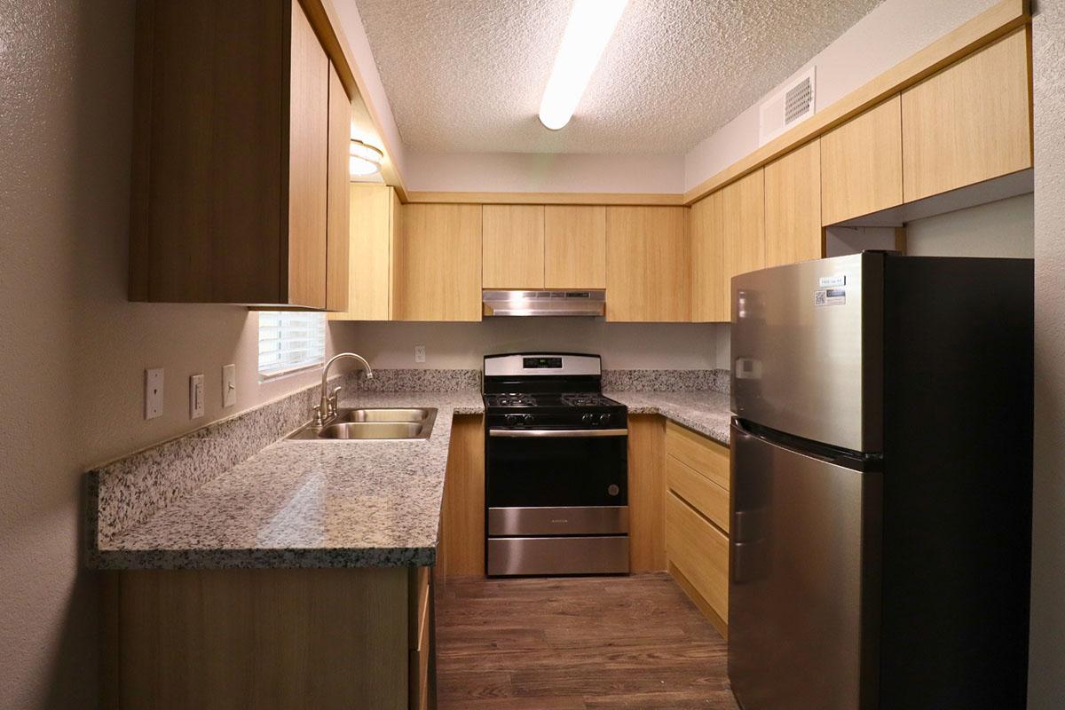 Modern kitchen with light wood cabinetry, granite countertops, a stainless steel sink, a gas stove, and a refrigerator. The space features overhead lighting and a window for natural light. The flooring is a warm, wood-like finish, creating a cozy atmosphere.