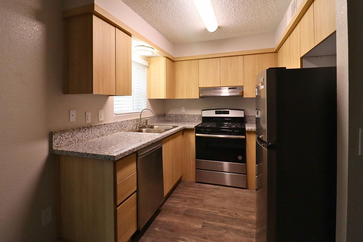 A modern kitchen featuring light wood cabinets, a stainless steel oven and stovetop, a black refrigerator, and a granite countertop. Natural light enters through a window above the sink, with a simple ceiling light providing illumination. The flooring is a dark wood-like material, creating a warm, inviting space.