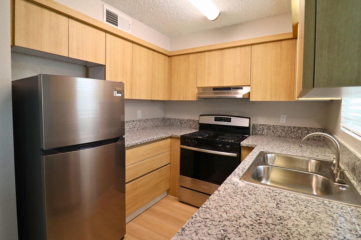 Modern kitchen featuring light wooden cabinetry, stainless steel refrigerator, and a gas stove with oven. The countertops are made of speckled granite, and a double sink is visible. The space is well-lit with overhead lighting and a window, creating a bright and functional cooking area.