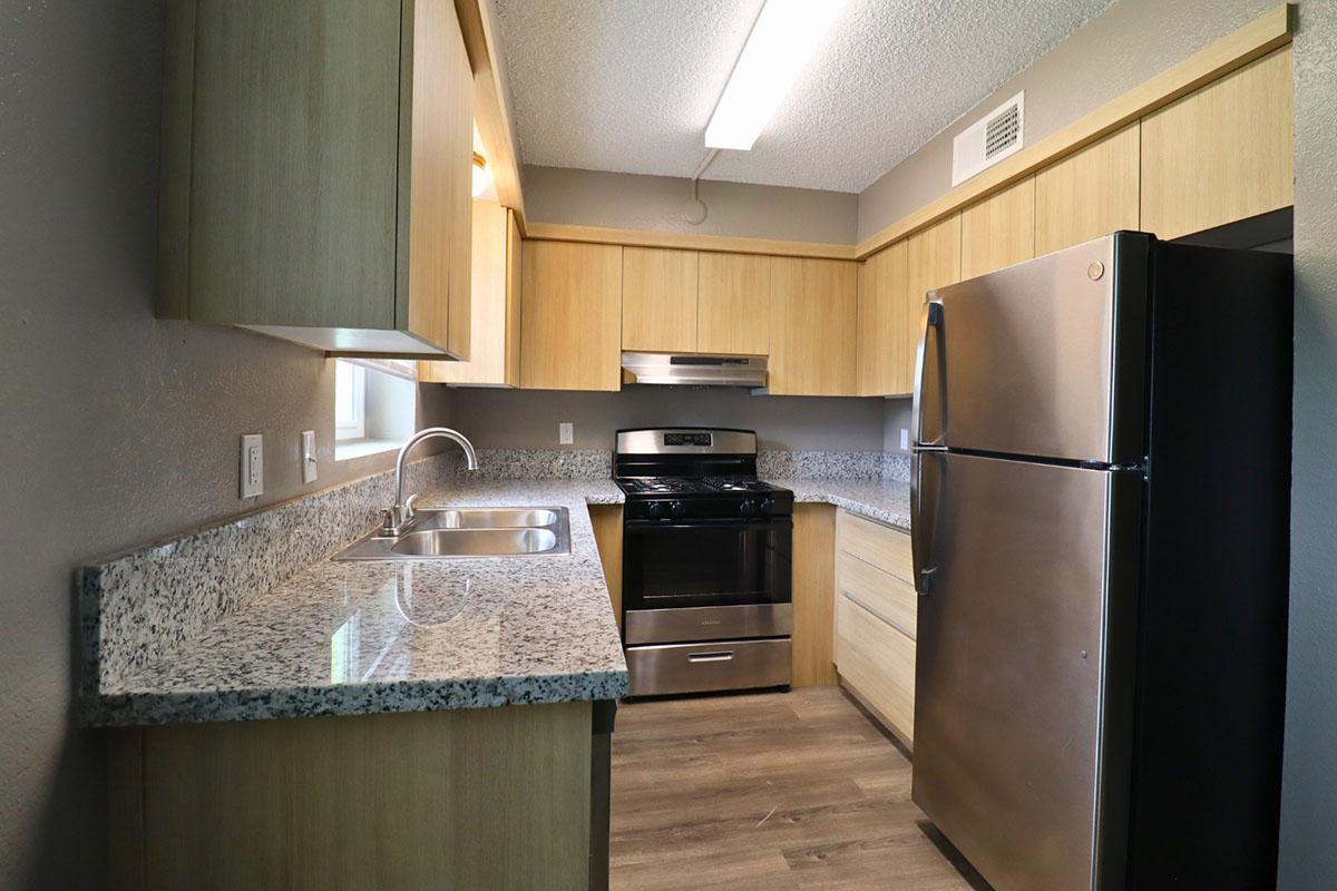 A modern kitchen featuring wooden cabinetry, granite countertops, a stainless steel refrigerator, a black oven and stove, and a double sink. The space is well-lit with overhead lighting and has a neutral color scheme, showcasing a clean and functional layout.