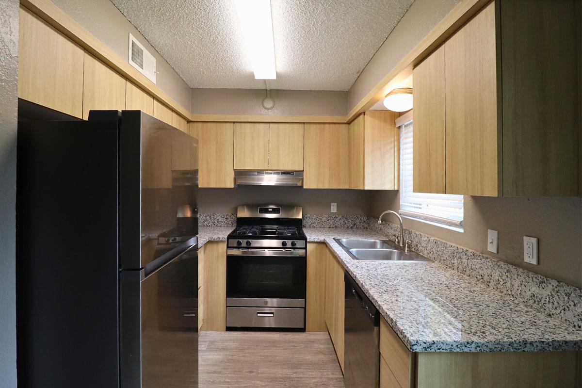 Modern kitchen featuring light wooden cabinetry, granite countertops, a stainless steel refrigerator, and a gas stove. The space is well-lit with overhead lighting and a window, showcasing a clean and functional design ideal for cooking and dining.