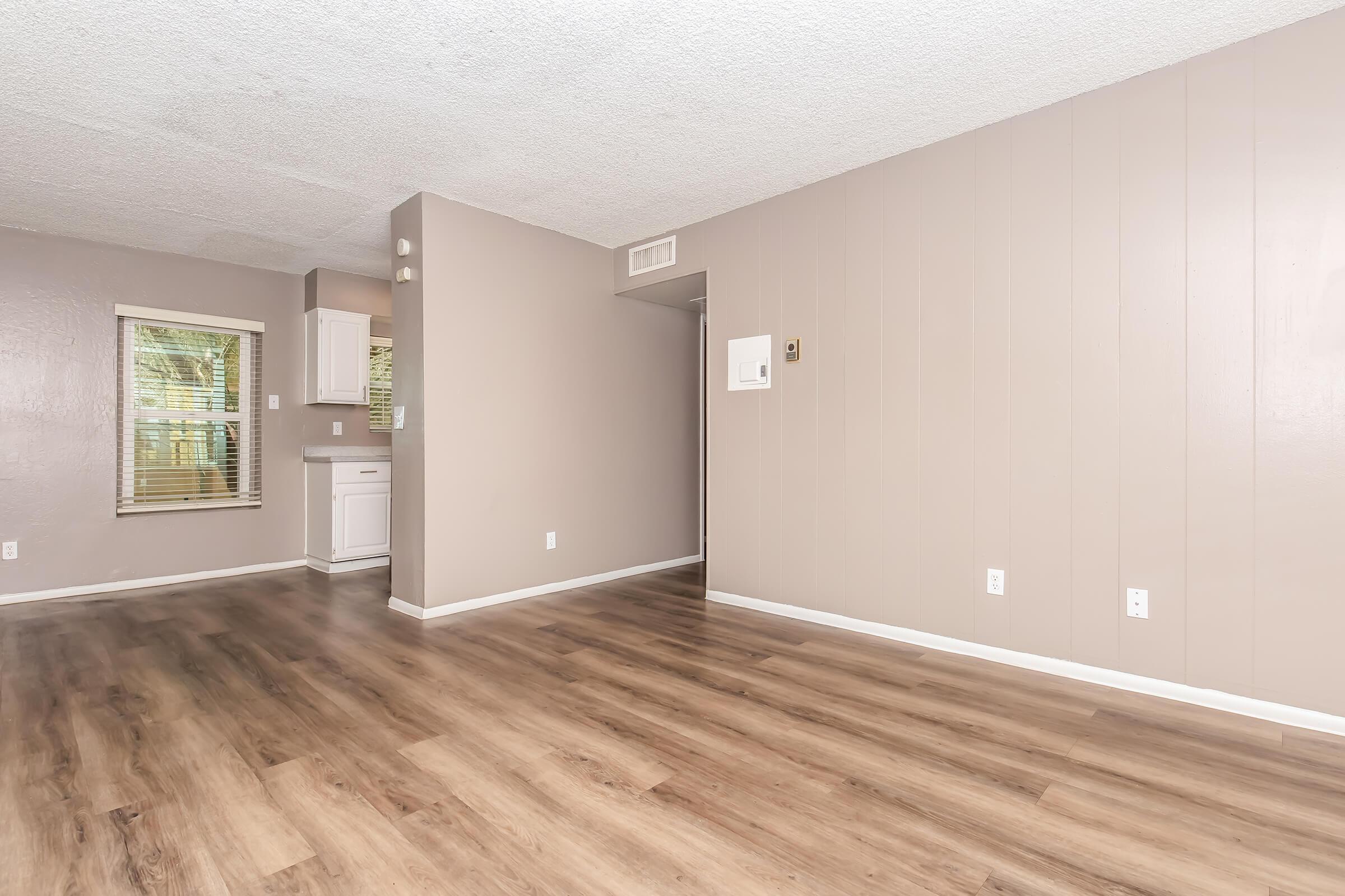 Spacious living area with wooden flooring, light gray walls, and a large window allowing natural light. A glimpse of a kitchen area with white cabinetry is visible in the background. The room is open and uncluttered, creating a welcoming atmosphere.