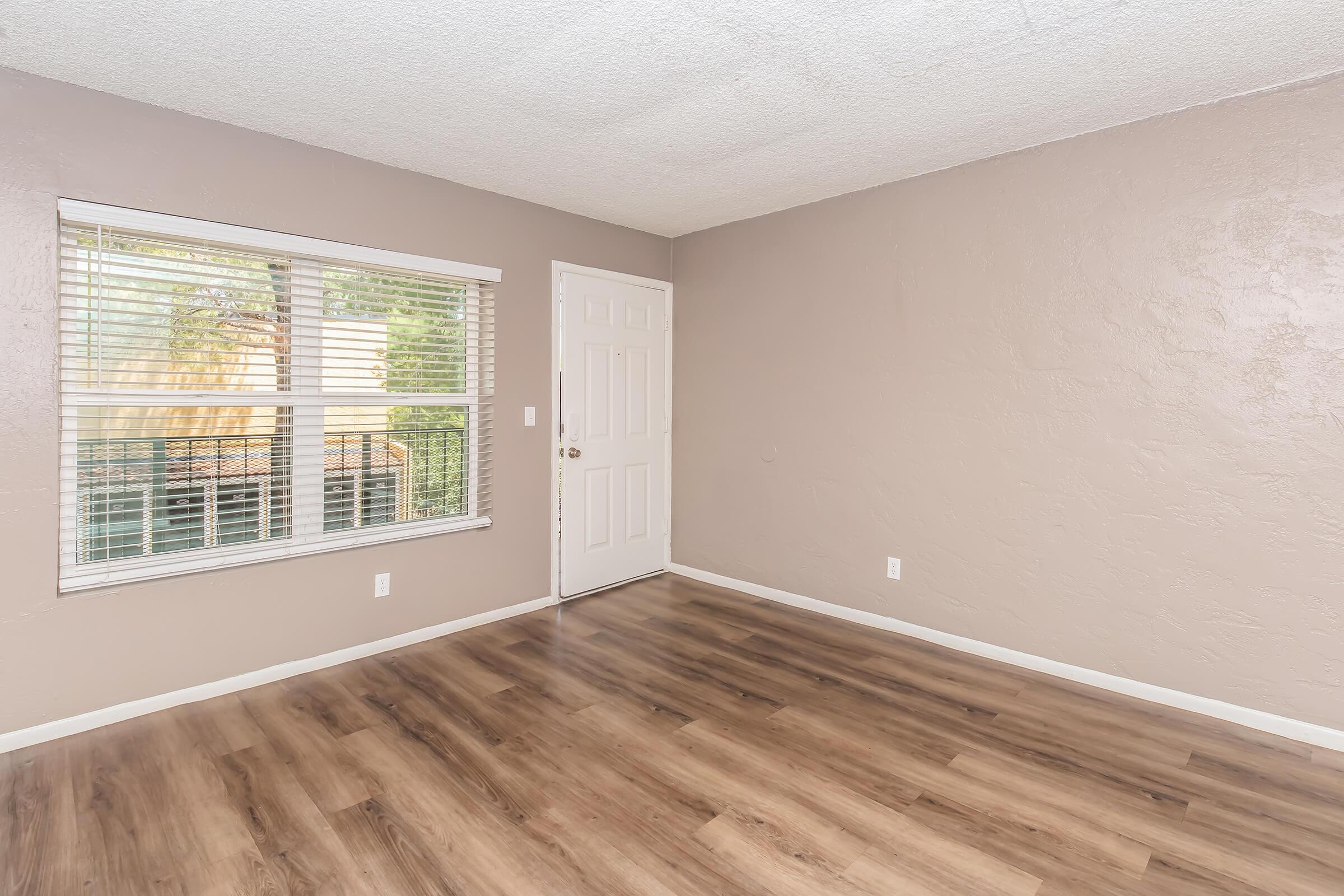 A spacious, empty room with light brown walls and laminate flooring. A large window with horizontal blinds allows natural light to filter in. The door is white, leading to the exterior, and the overall ambiance is bright and inviting. The room is ready for furniture and personal touches.