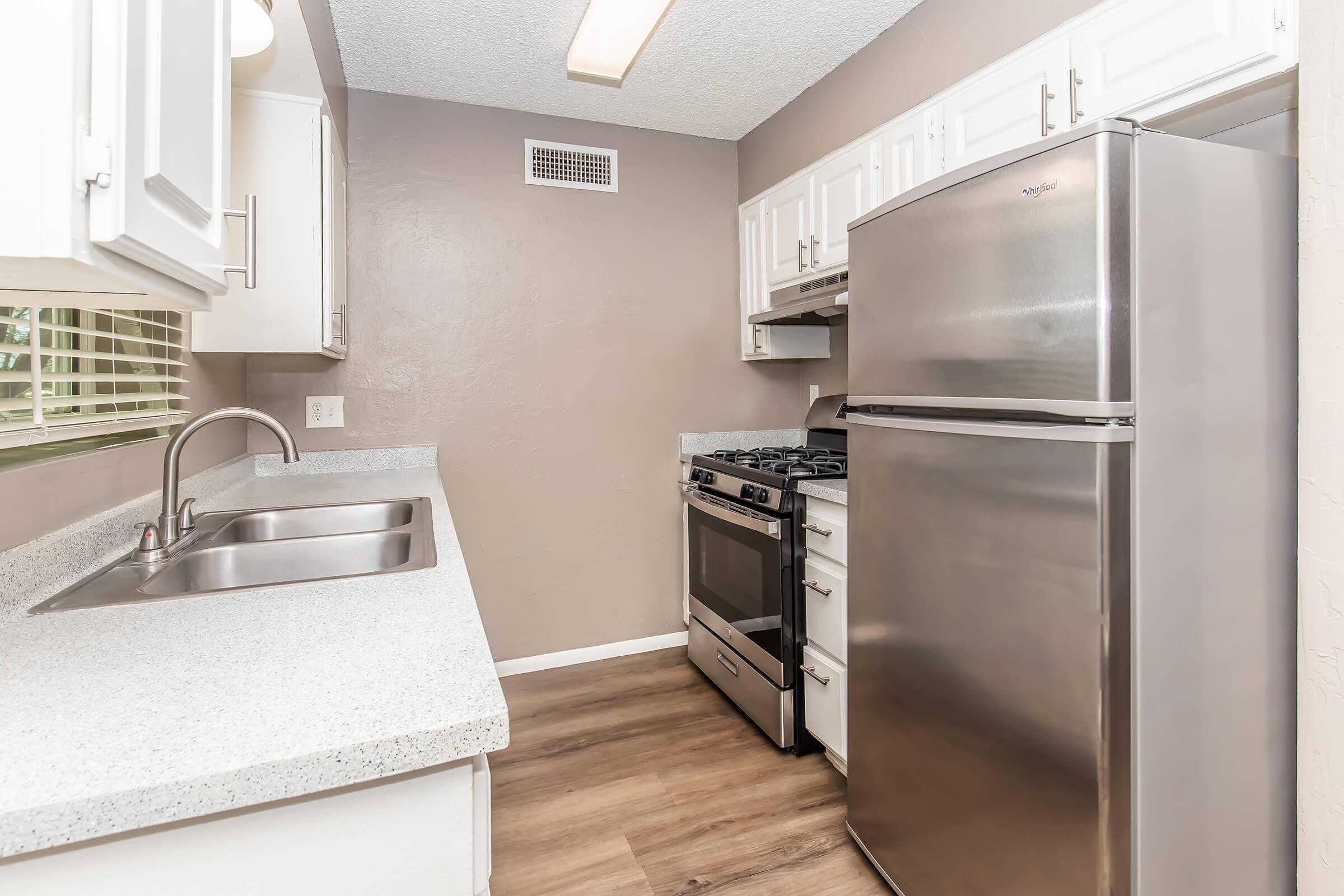 A modern kitchen featuring stainless steel appliances including a refrigerator and a gas stove, white cabinetry, and a sink. The countertop is a light gray material, and the walls are painted a soft neutral color. Ample natural light is provided by a window. The flooring is a warm wood look.