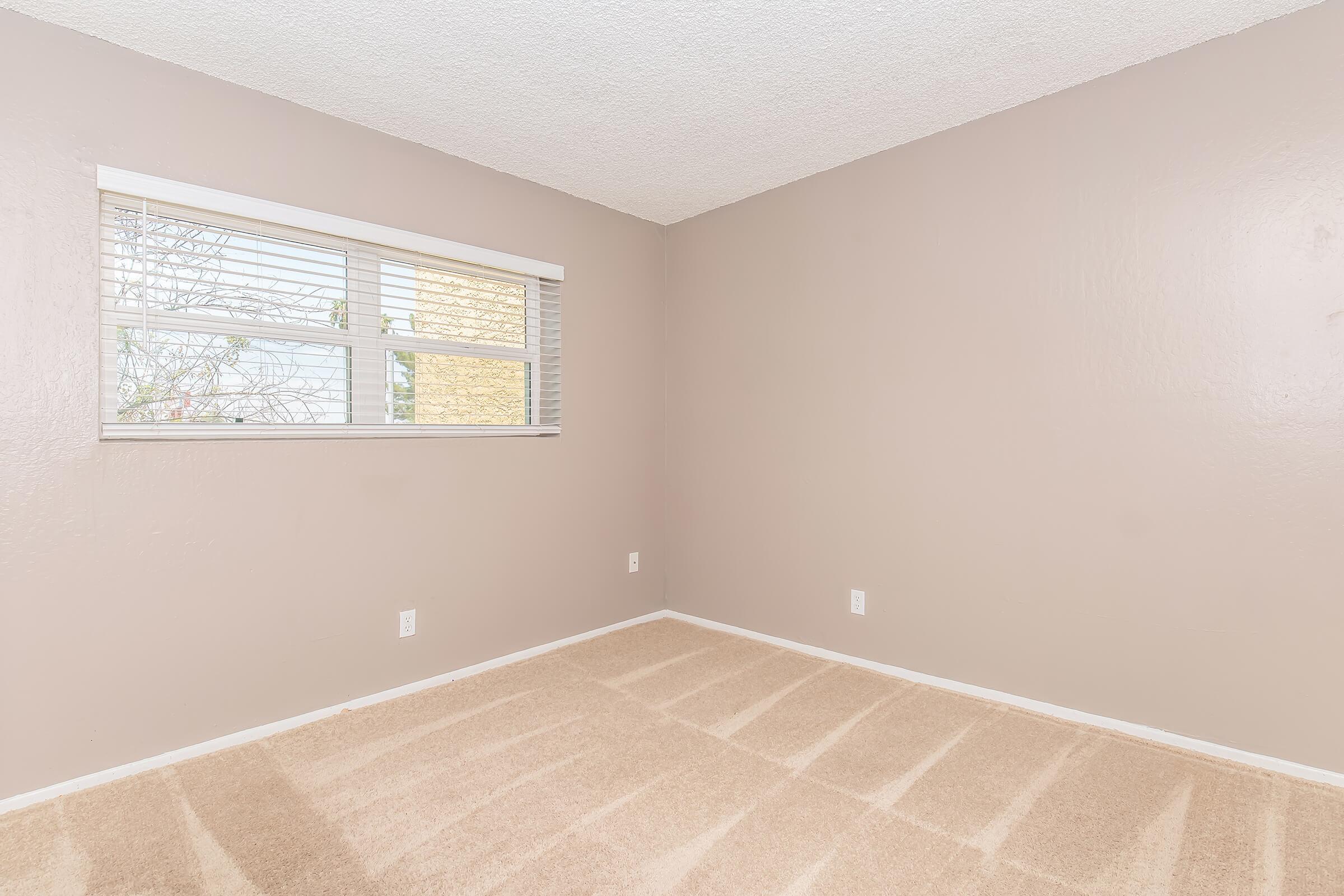 A clean, empty room with beige walls and carpet. There's a window with blinds on one wall, allowing natural light to enter. The space appears simple and well-maintained, ideal for various uses. The floor shows light patterns, possibly from the window's light.
