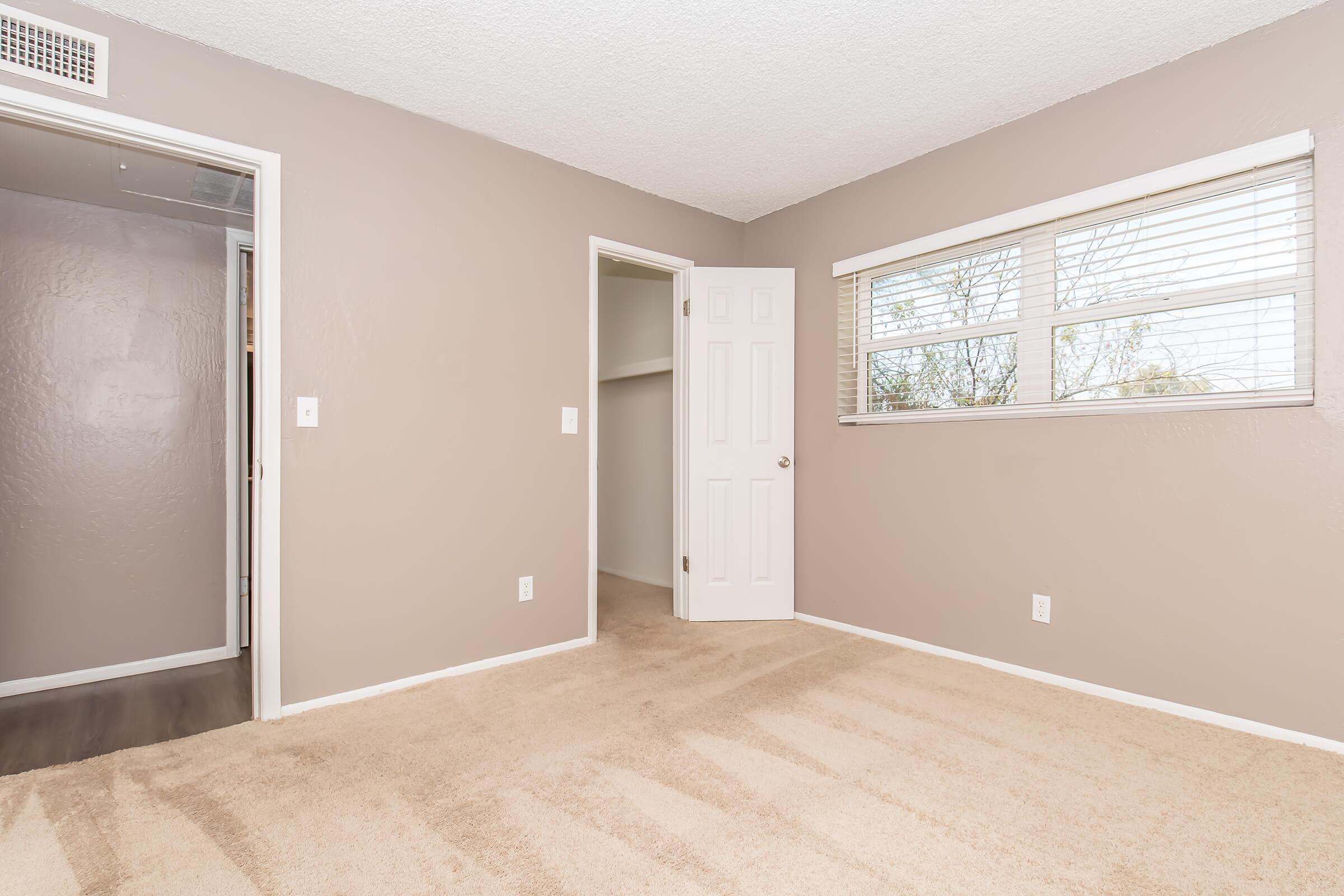 A well-lit, empty room with beige carpet and light brown walls. There are two doors: one leading to a closet and the other to a hallway. A window with blinds allows natural light to enter. The space appears ready for decoration or furnishing.