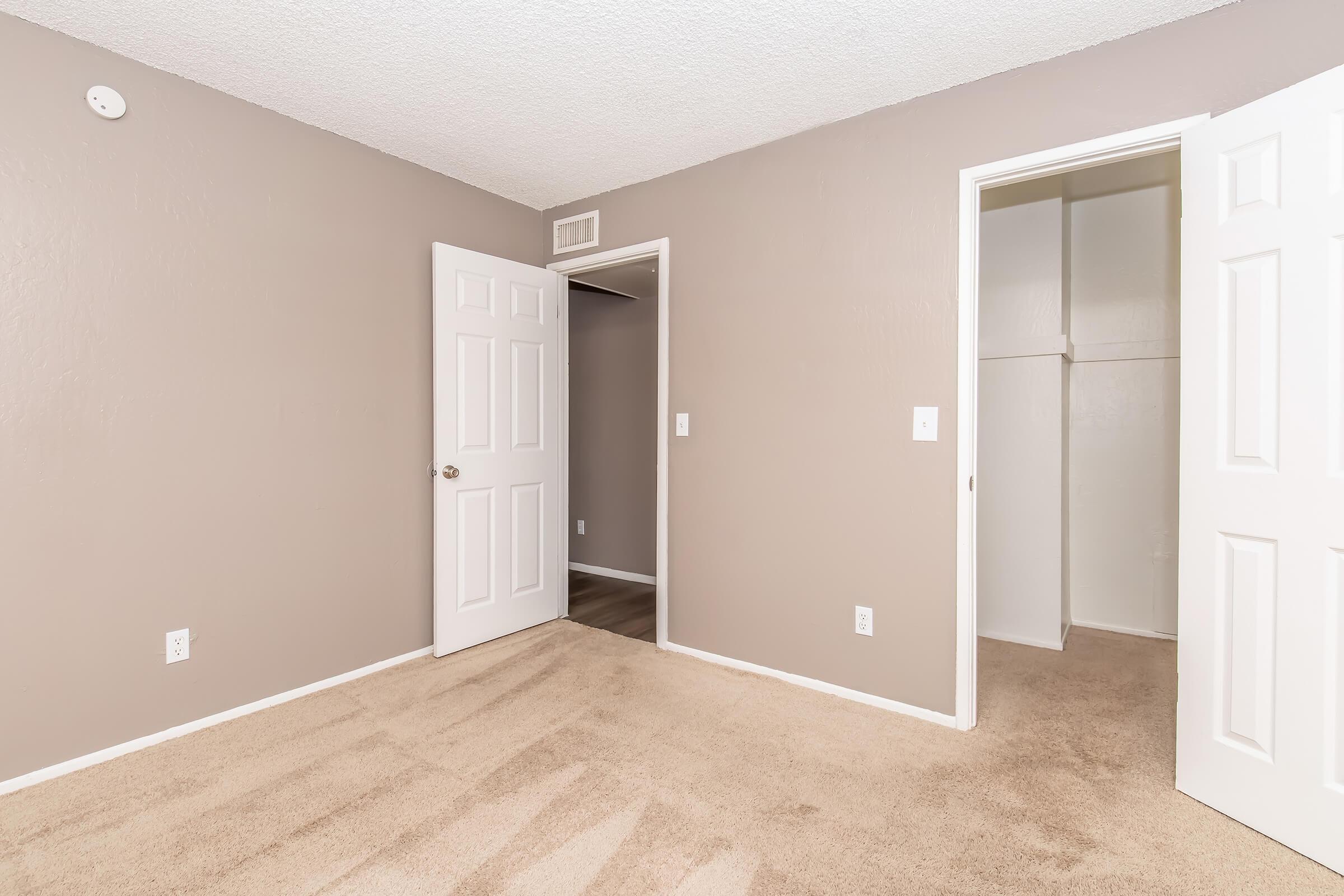 A beige room with textured walls, featuring two white doors. One door leads to a closet, while the other opens into a hallway. The floor is covered with light brown carpet, and there is a small vent on the left wall. Natural light enters through the open doors, creating a bright and airy atmosphere.