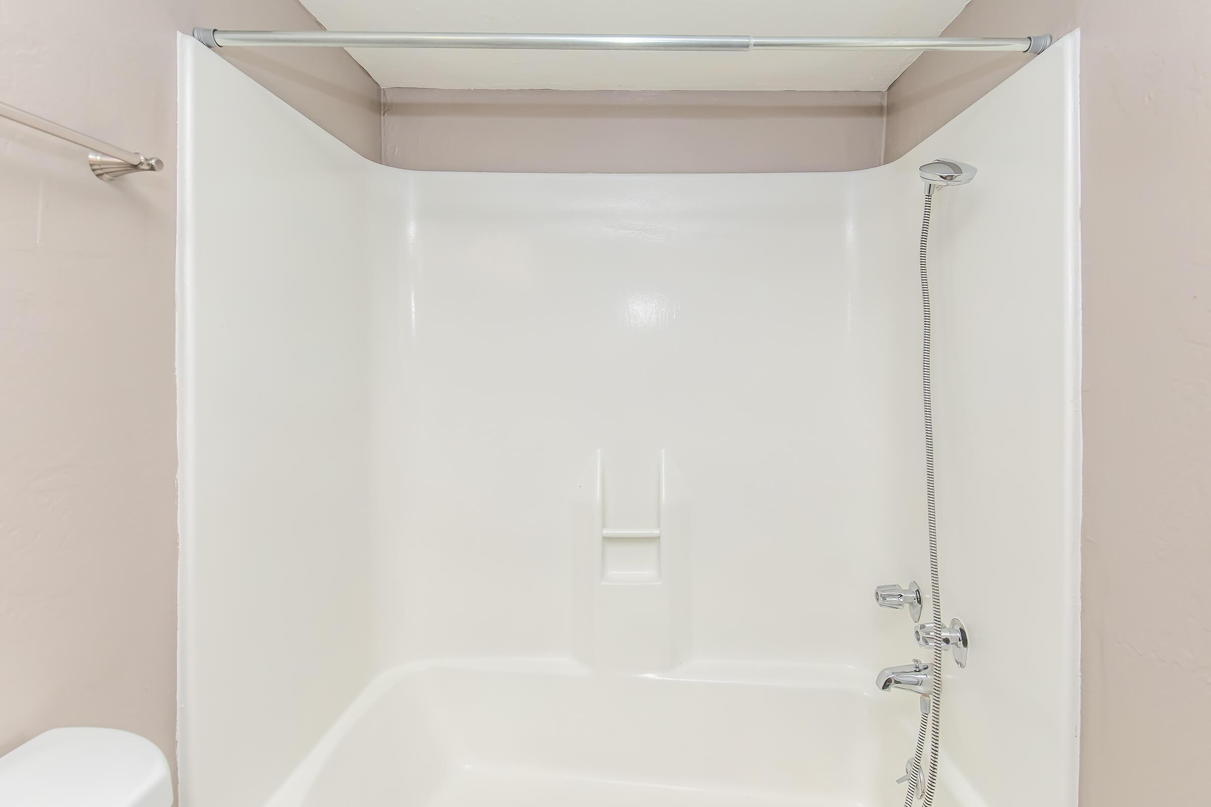 A clean, empty shower area with white walls and a silver showerhead. The shower features a built-in shelf for toiletries and a straightforward design. The background is a light, neutral color, creating a bright and spacious appearance.