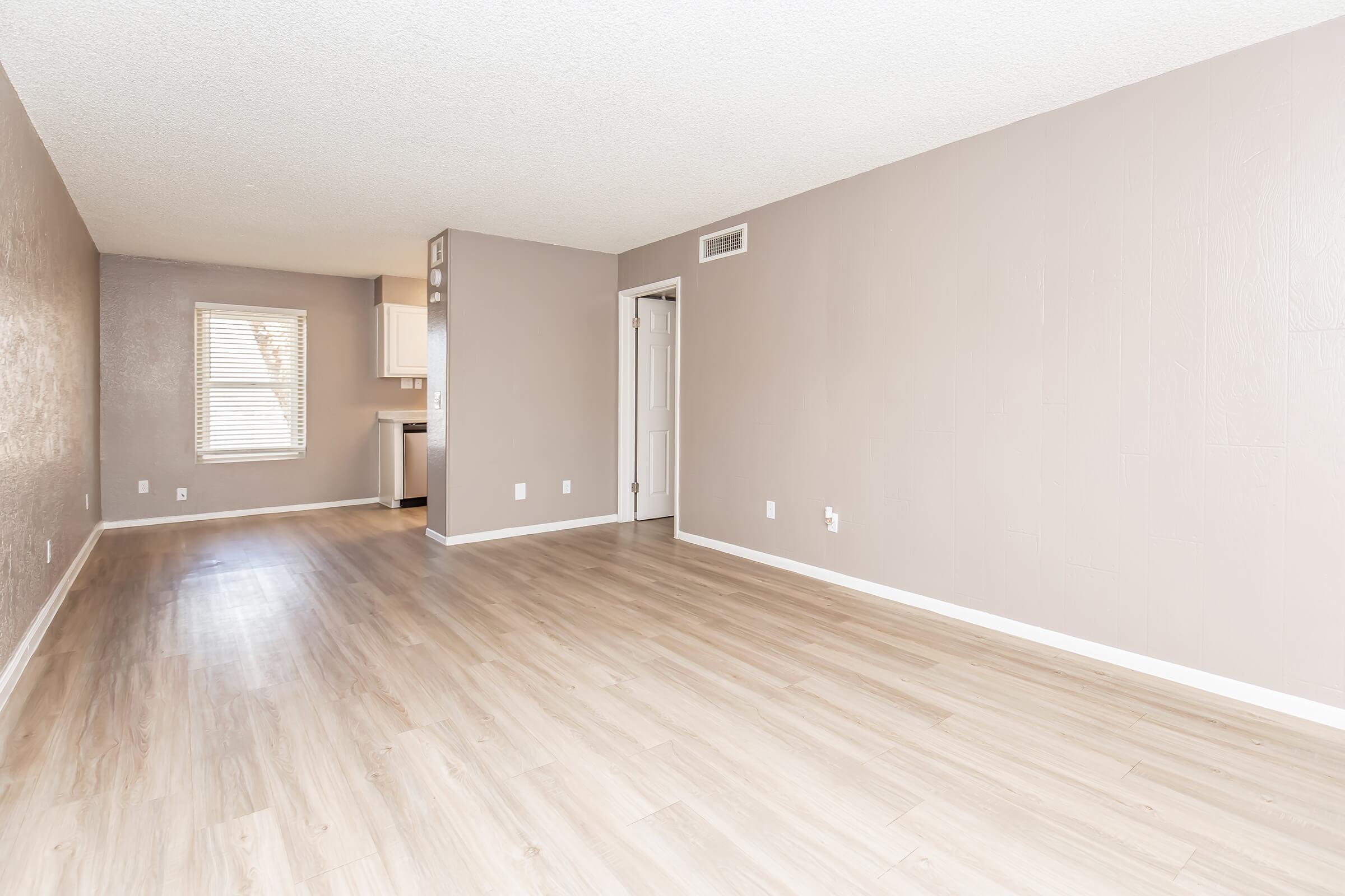 A spacious, light-filled room with light wood-like flooring and neutral beige walls. A window allows natural light to enter, and a doorway leads to another area, possibly a kitchen. The room is empty, offering a blank canvas for furnishings and decor.