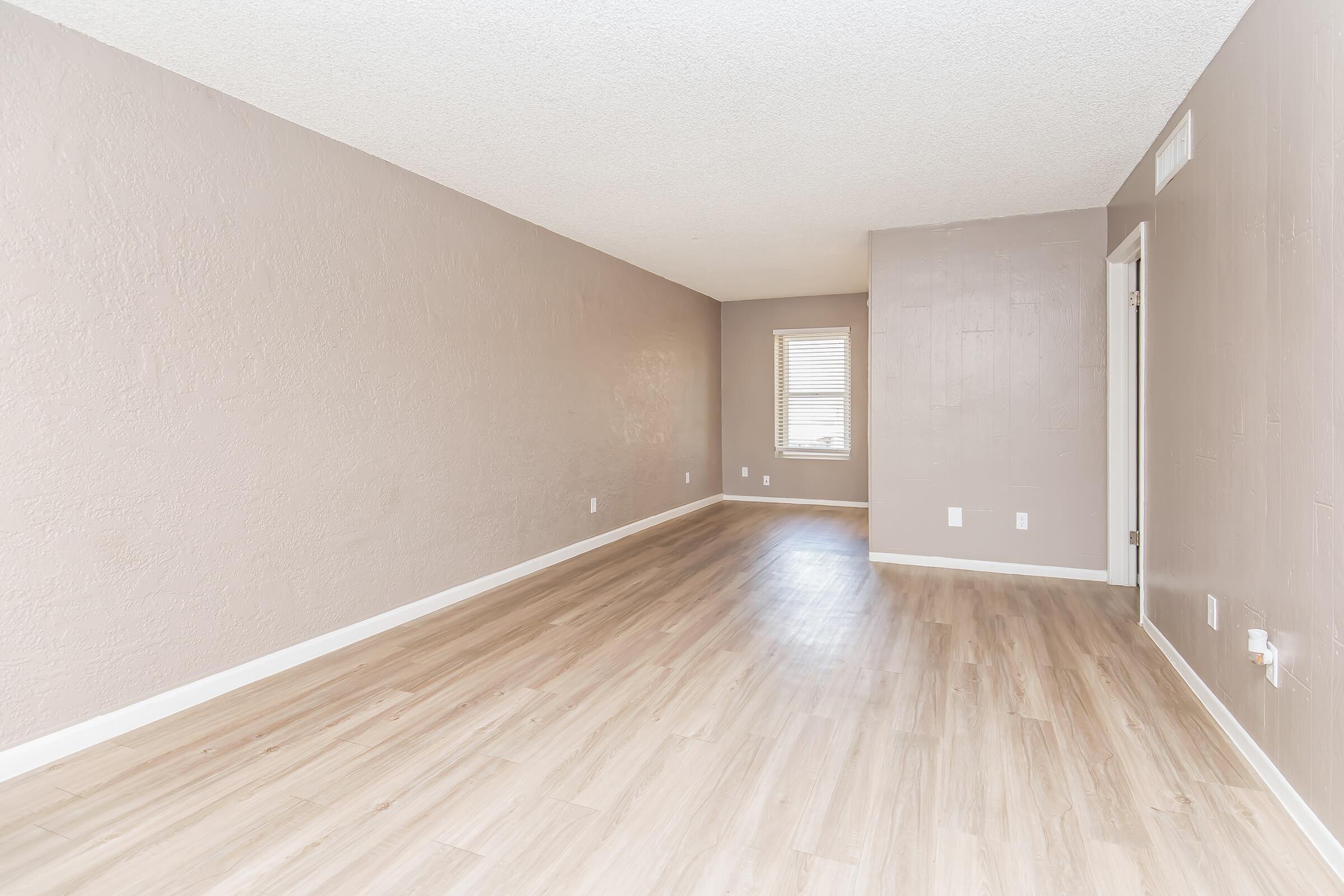 A spacious, empty room with light brown walls and wooden flooring. A single window lets in natural light, creating a bright atmosphere. The room features smooth walls and minimal furnishings, making it ideal for various design options. There are doors leading to adjacent spaces.