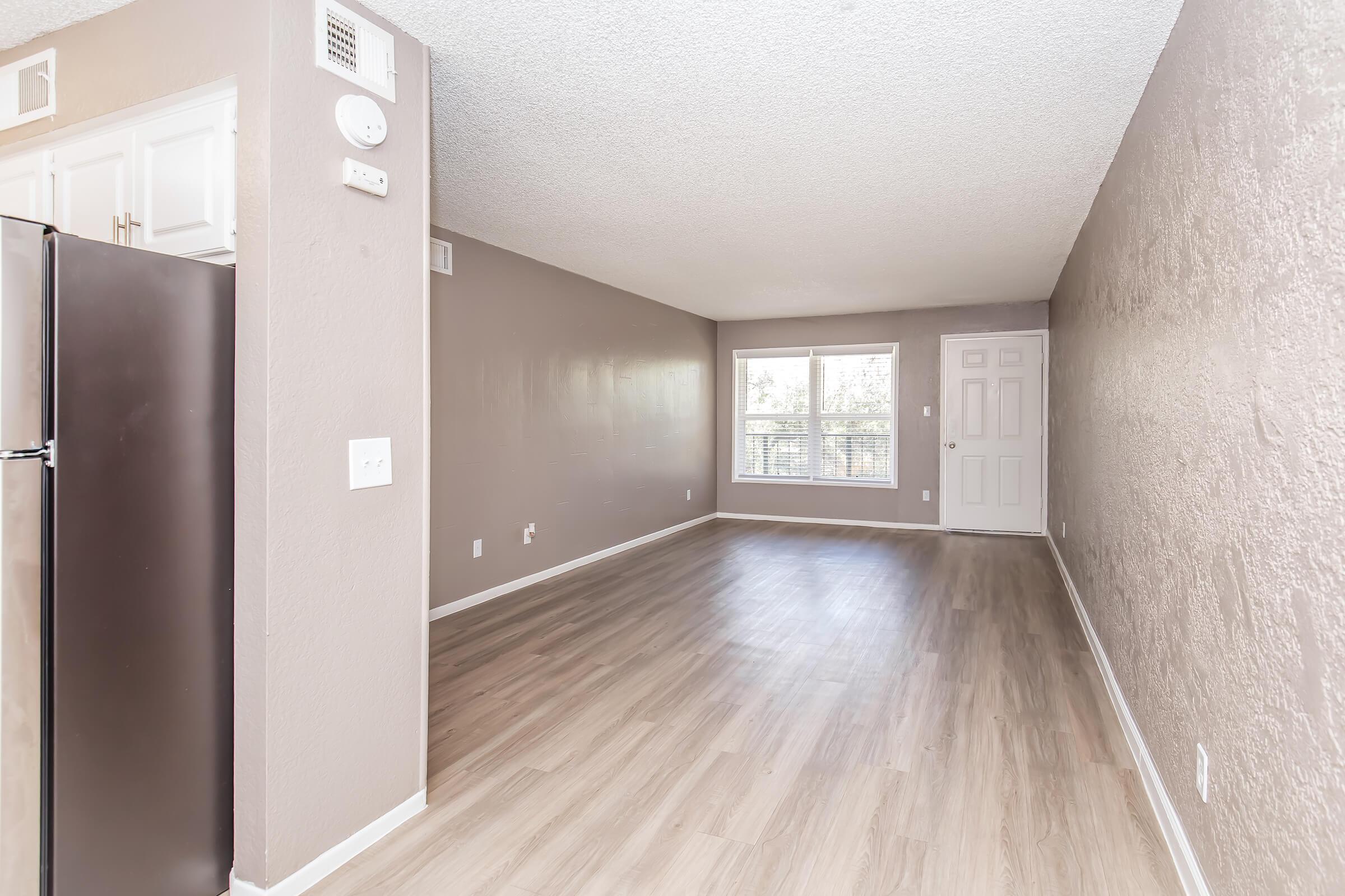 A spacious, empty living area with light-colored walls and a textured ceiling. There is a window allowing natural light, a beige floor, and a kitchen area featuring a black refrigerator. The room appears well-lit and ready for decoration.