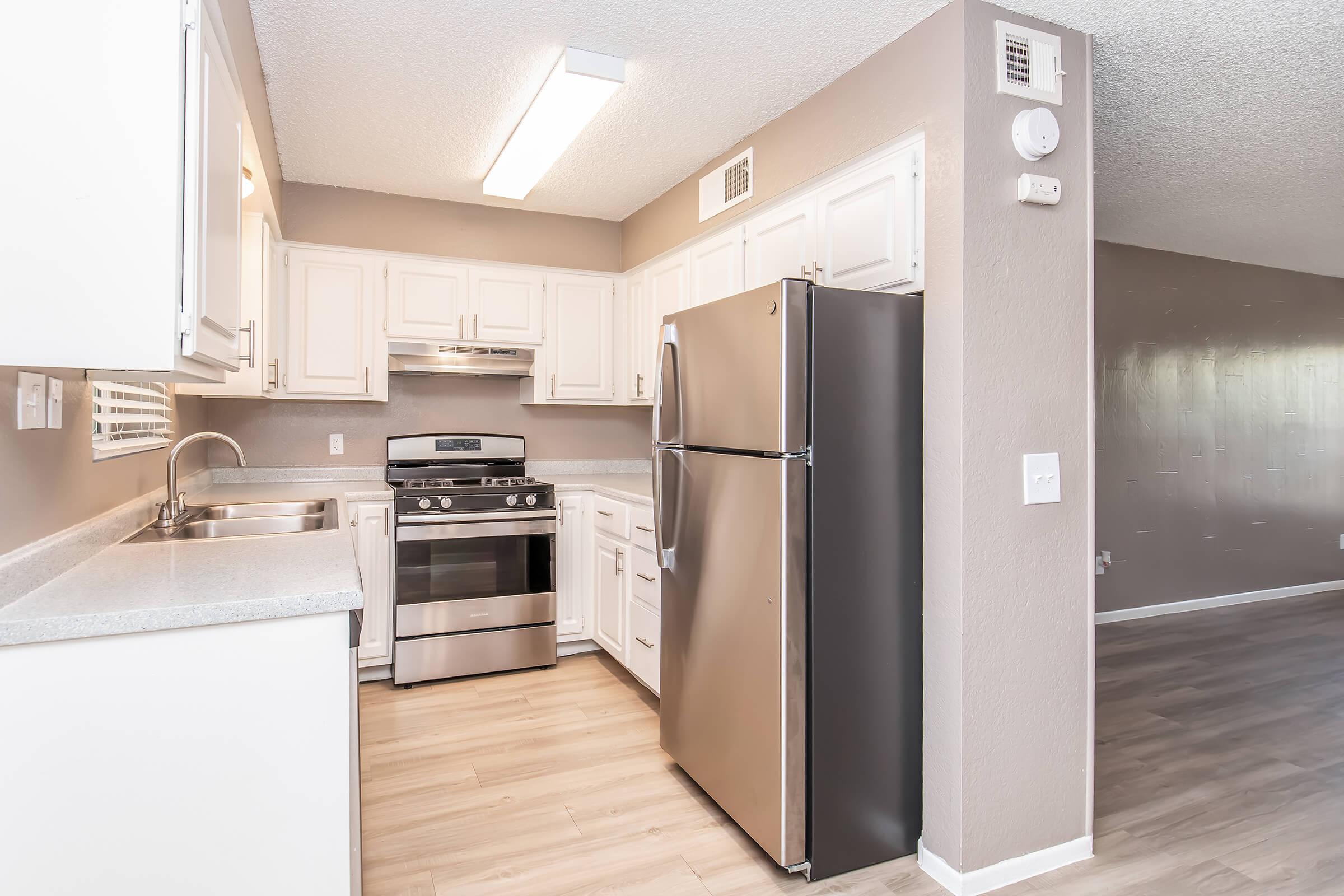 A modern kitchen featuring white cabinetry, a stainless steel refrigerator, and a stainless steel gas stove. The countertops are light gray, and the flooring is a light wood laminate. Bright overhead lighting enhances the spacious feel of the kitchen, with a neutral wall color complementing the design.