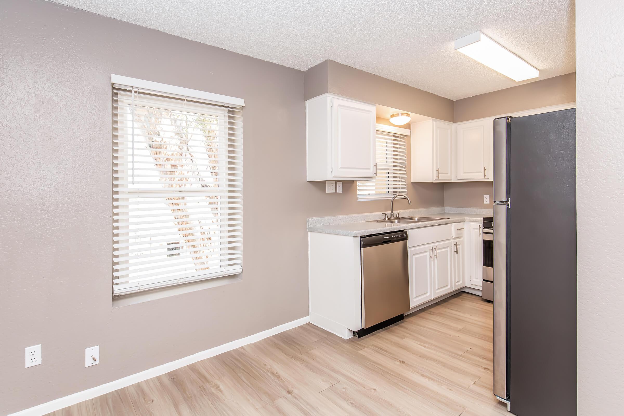 Modern kitchen featuring white cabinetry, stainless steel appliances, and grey walls. A window with blinds allows natural light, illuminating the countertop and sink area. The flooring is light-colored, enhancing the overall brightness of the space.