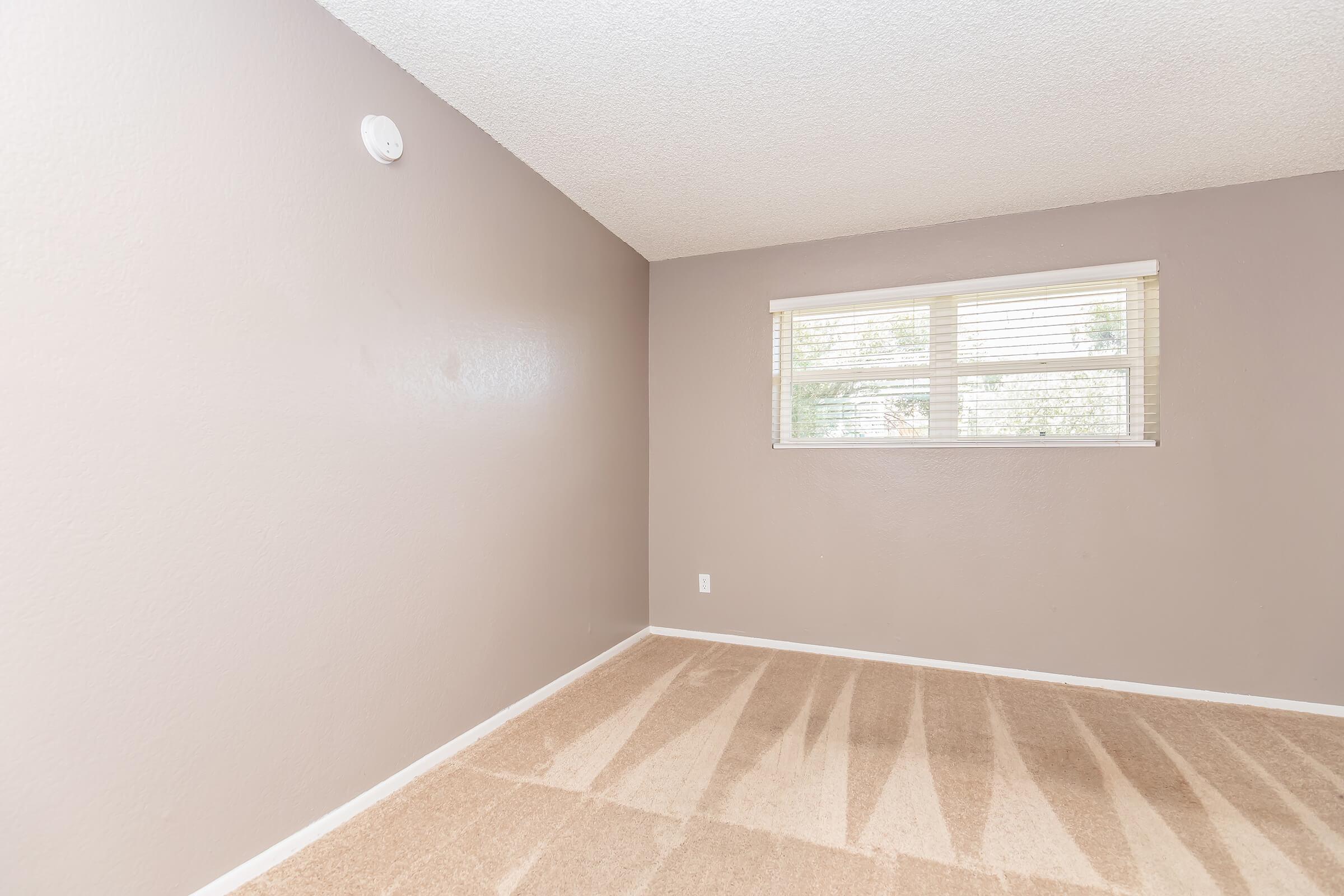 A vacant room with light brown walls and beige carpet. There is a window with blinds on one wall, allowing natural light to enter. The room appears tidy and unadorned, with a smoke detector mounted on the wall near the ceiling.