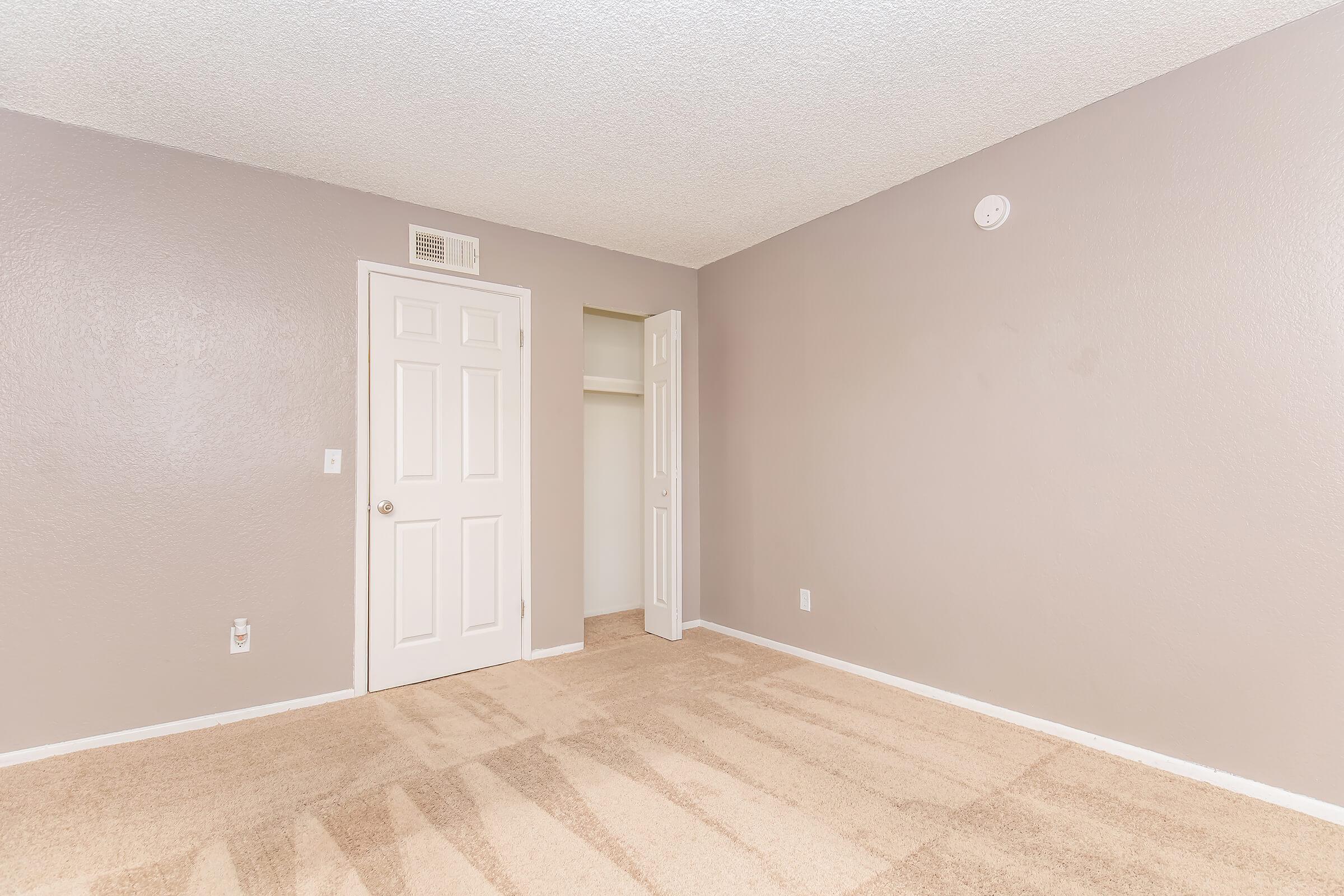 An empty room with light gray walls and beige carpet. There's a closed white door on the left and an open door leading to a closet on the right. The room is well-lit, displaying a clean and spacious environment. A small round fixture is visible on the ceiling.