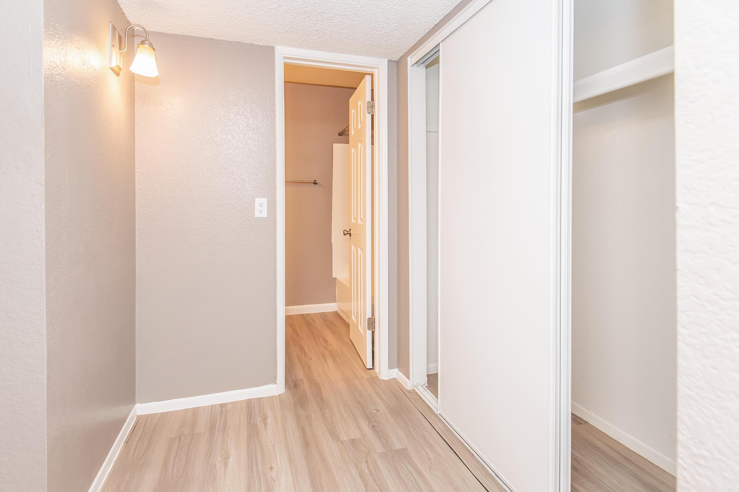 A well-lit room with light gray walls and new laminate flooring. There is an open door leading to a small closet space, which features a sliding door. The room is enhanced by wall-mounted lighting, creating a bright and inviting atmosphere.