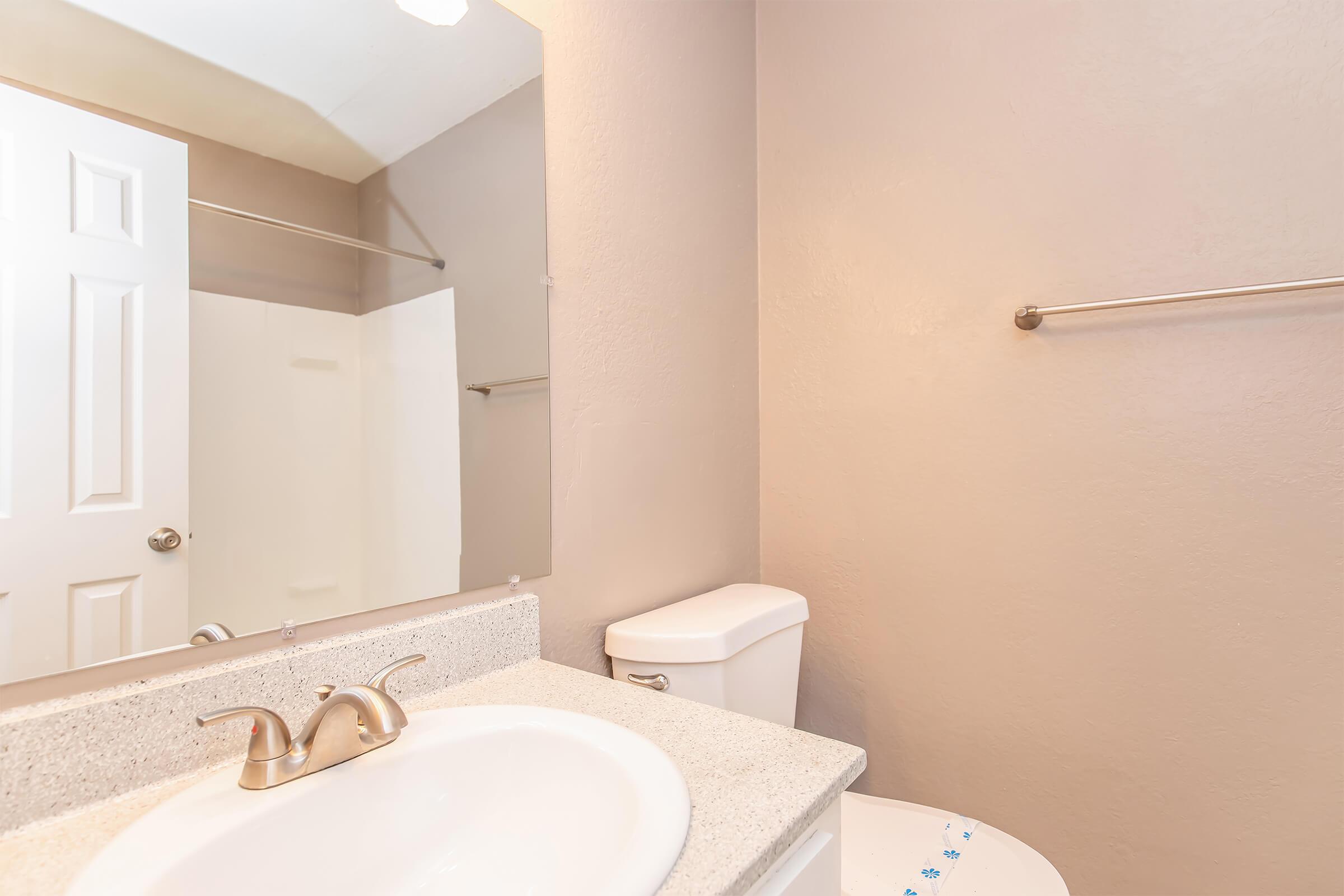 A clean and modern bathroom featuring a white toilet, a sink with a granite countertop, and a large mirror. The walls are painted a light beige color, and a shower area is visible behind a sliding door. A towel rack is installed beside the sink.