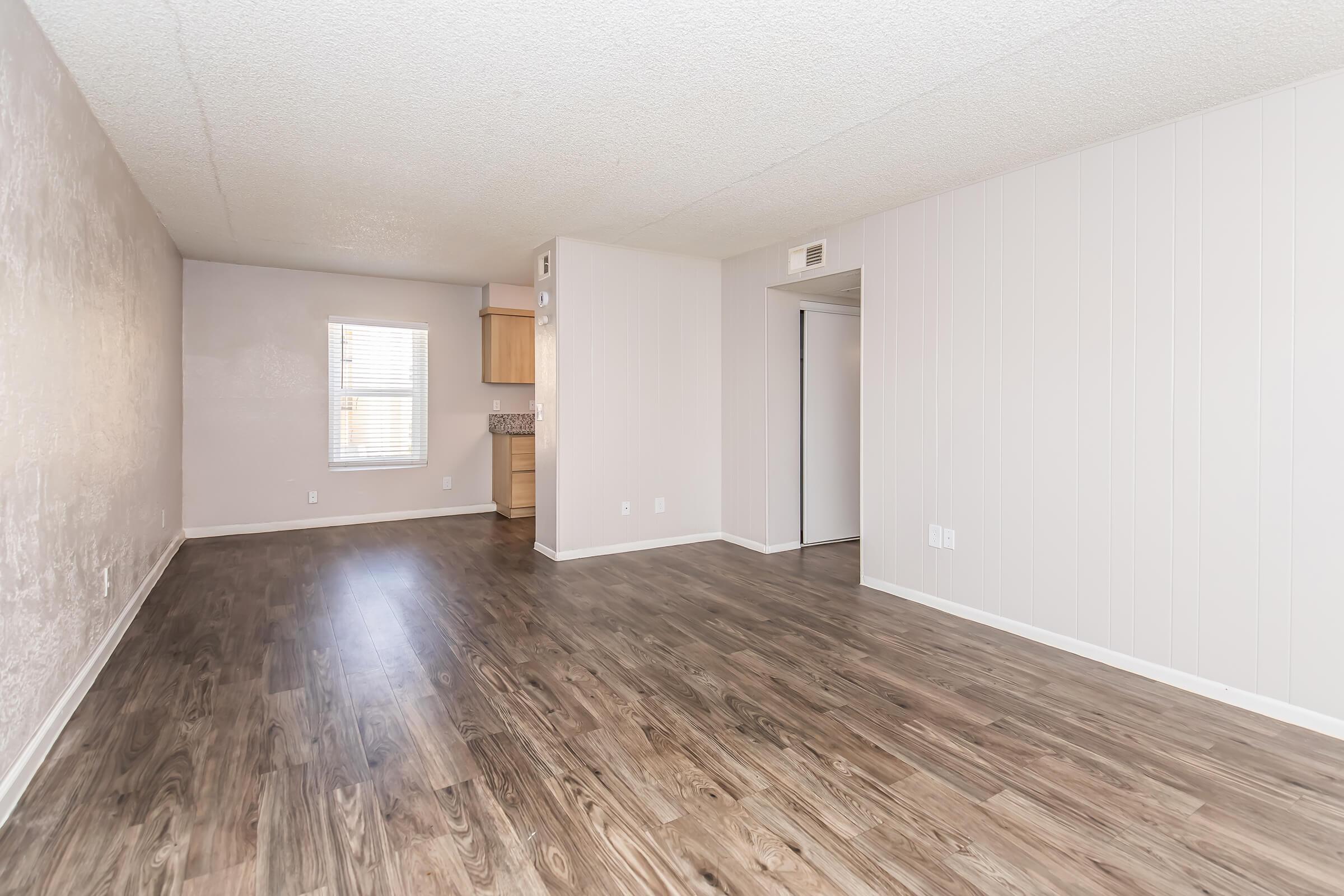 A spacious, empty living area with light-colored walls and laminate flooring. A window provides natural light, and a small kitchenette is visible in the background. The overall atmosphere is bright and ready for decoration or furnishing.