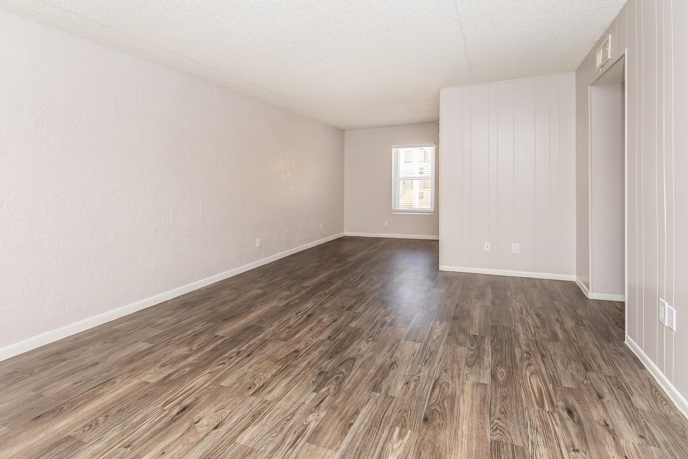 A spacious, empty room featuring light-colored walls and a wooden floor. Natural light enters through a window, illuminating the clear space. There's a doorway on the right leading to another area, and the overall ambiance is bright and neutral, suitable for various interior designs.