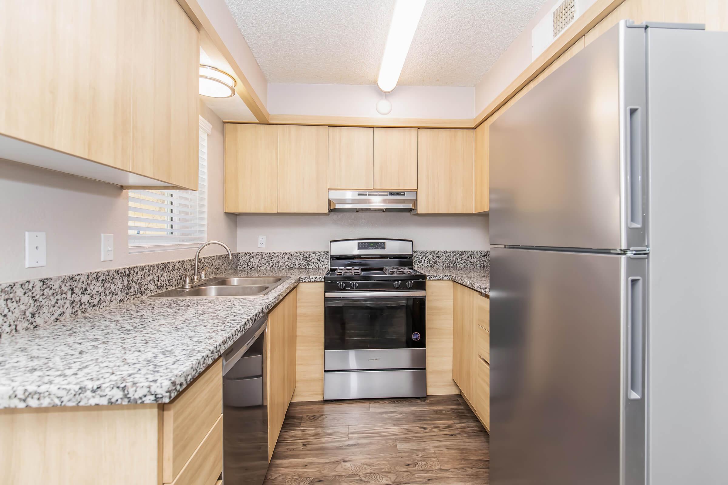 Modern kitchen featuring light wood cabinetry, granite countertops, a stainless steel refrigerator, and a gas stove. The space is well-lit with overhead lighting, and has a window allowing natural light. The floor is finished with a dark wood-like surface, creating a warm and inviting atmosphere.