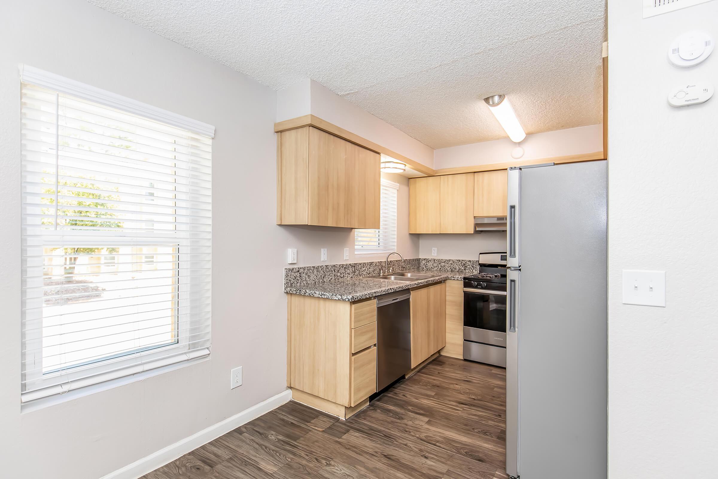 Modern kitchen featuring light wood cabinets, granite countertops, stainless steel appliances including a refrigerator and stove, and a sink. Large window with blinds provides natural light. Floor is a contemporary wood laminate.