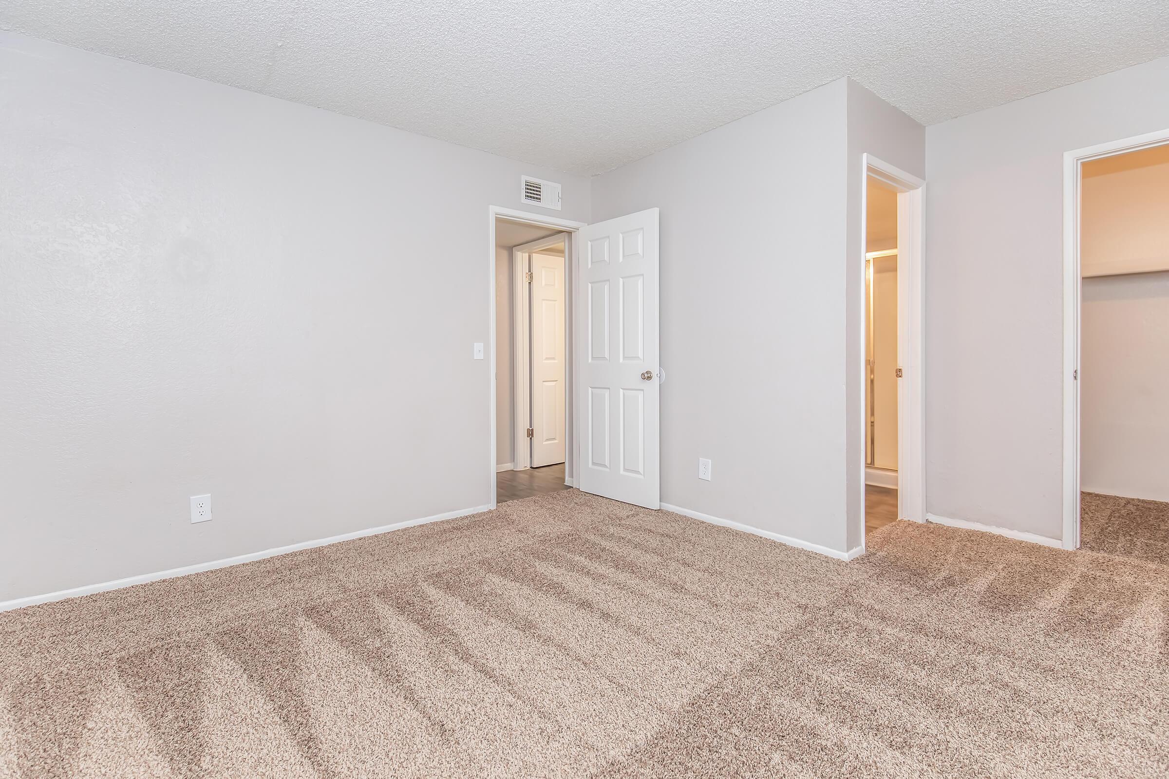 Spacious room with light gray walls and freshly carpeted floor. Two doorways lead to adjacent spaces, one featuring an open closet. The room is well-lit, with a textured ceiling enhancing the overall brightness. Ideal for an apartment or home interior setting.